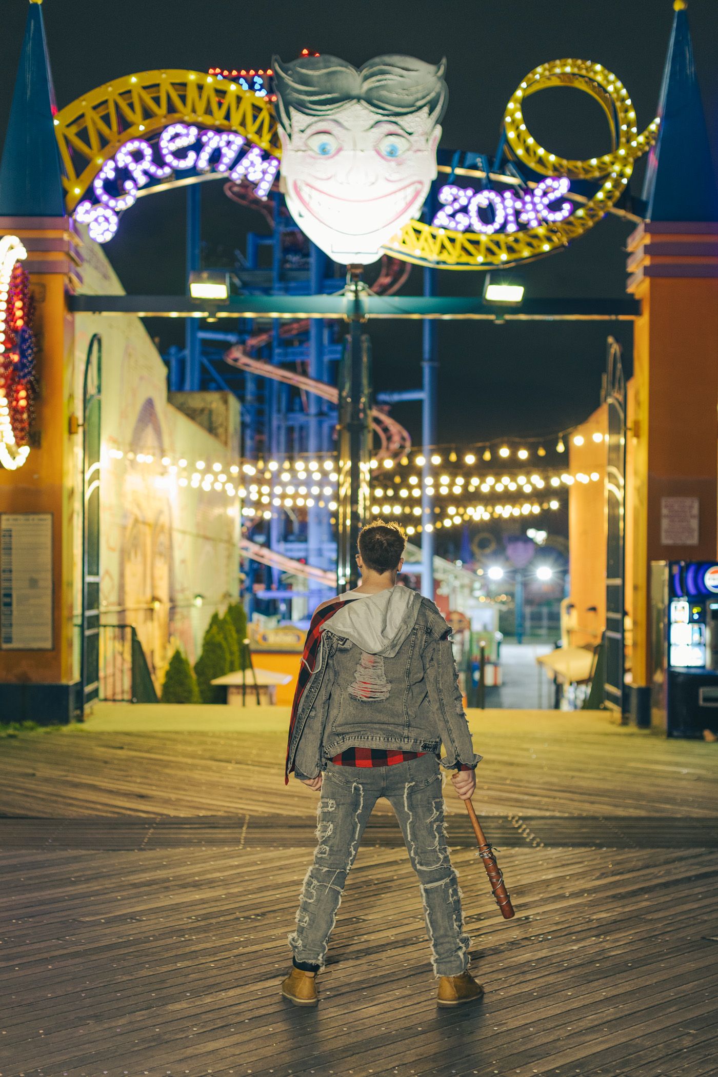 Person holding a baseball bat stands on a boardwalk at night facing a glowing Scream Zone amusement park sign with a giant smiling face.