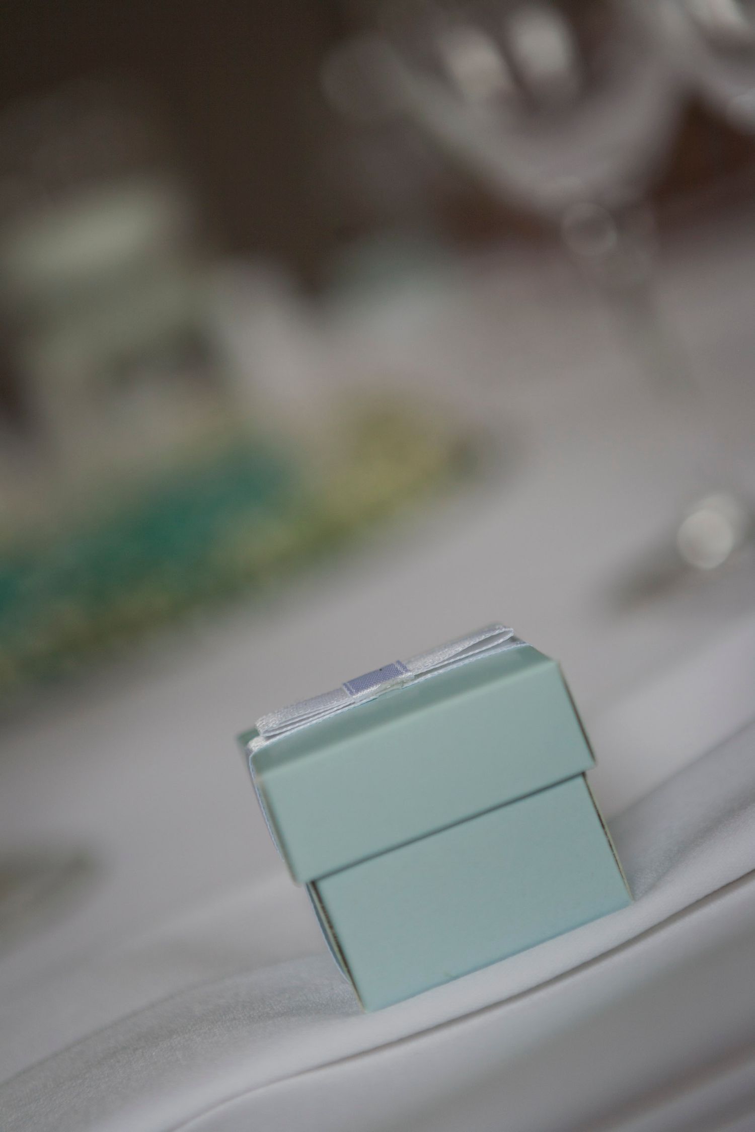 Teal wedding favor box on white tablecloth with blurred glassware in the background.