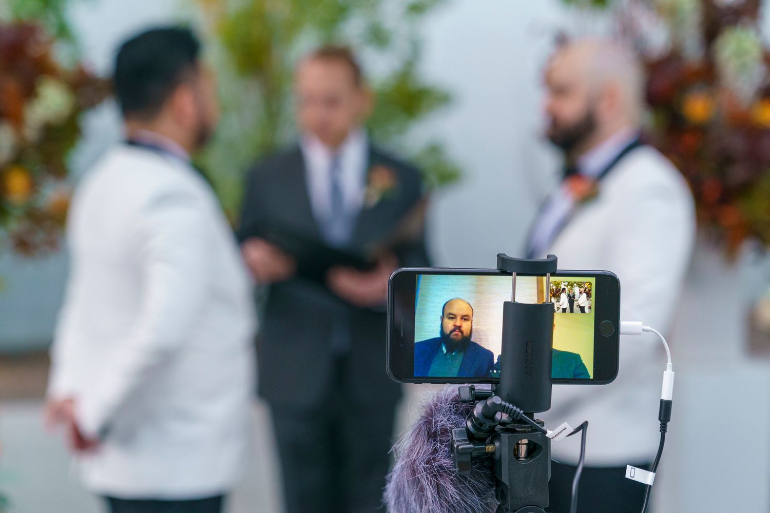 Same-sex wedding ceremony filmed on smartphone, two grooms in white suits with officiant in background.