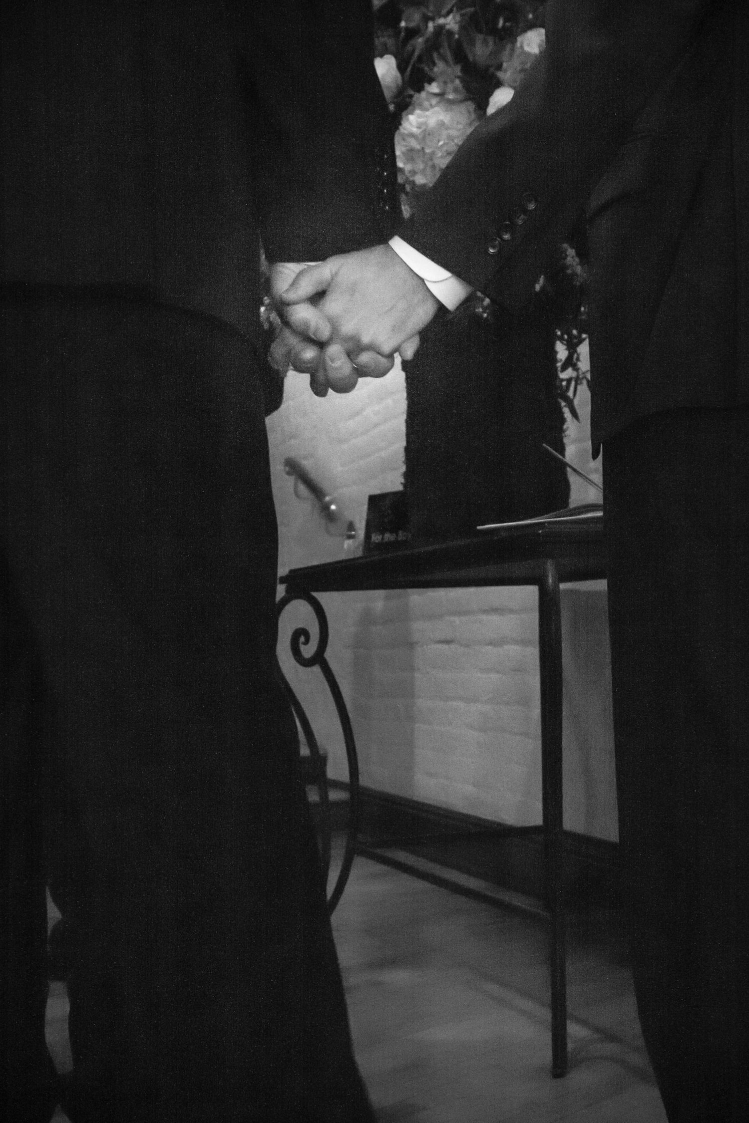 Black and white photo of two people in formal attire holding hands at a wedding ceremony near a decorative table.