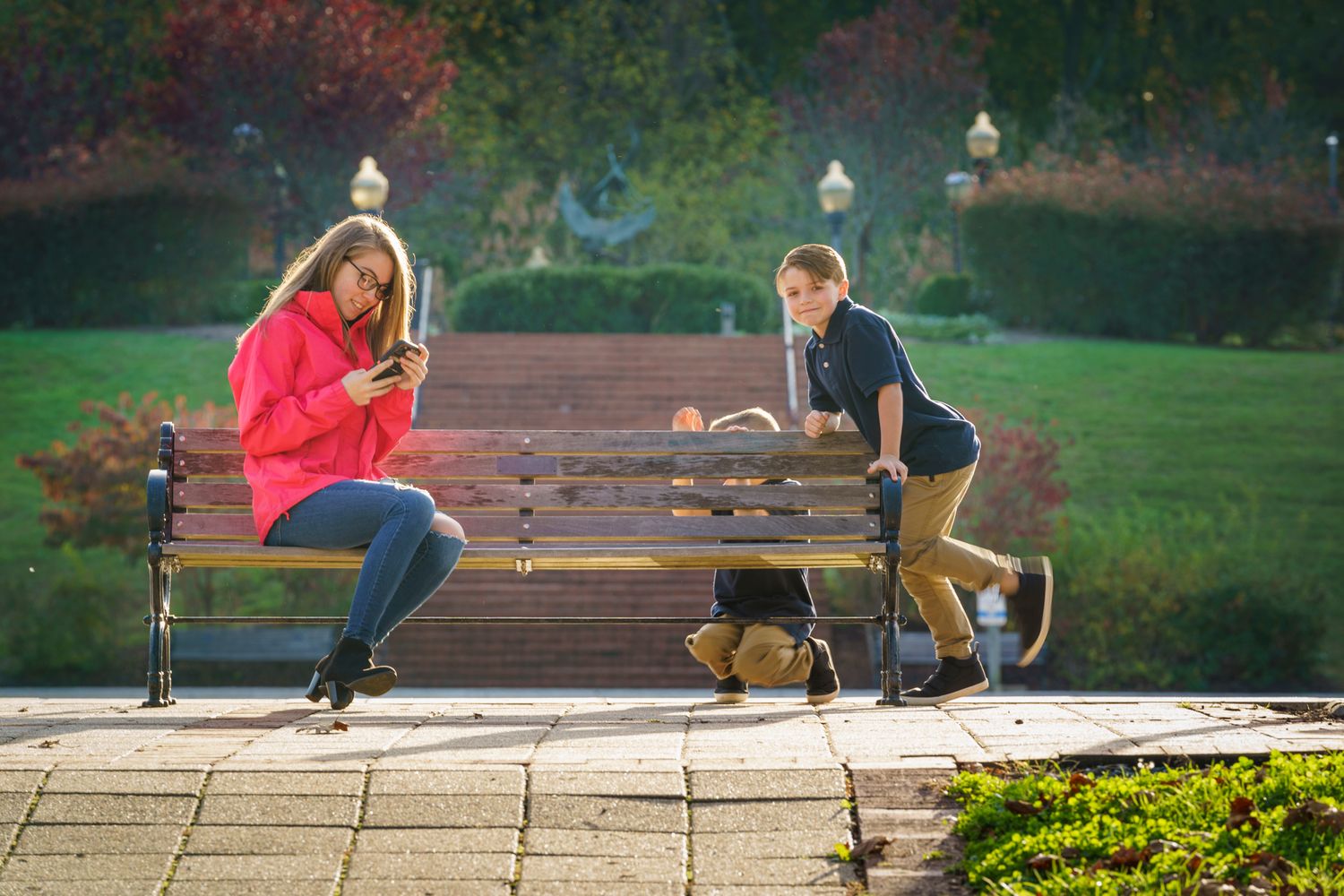 Woman in red jacket using phone on park bench while boy plays with dog on sunny autumn day.