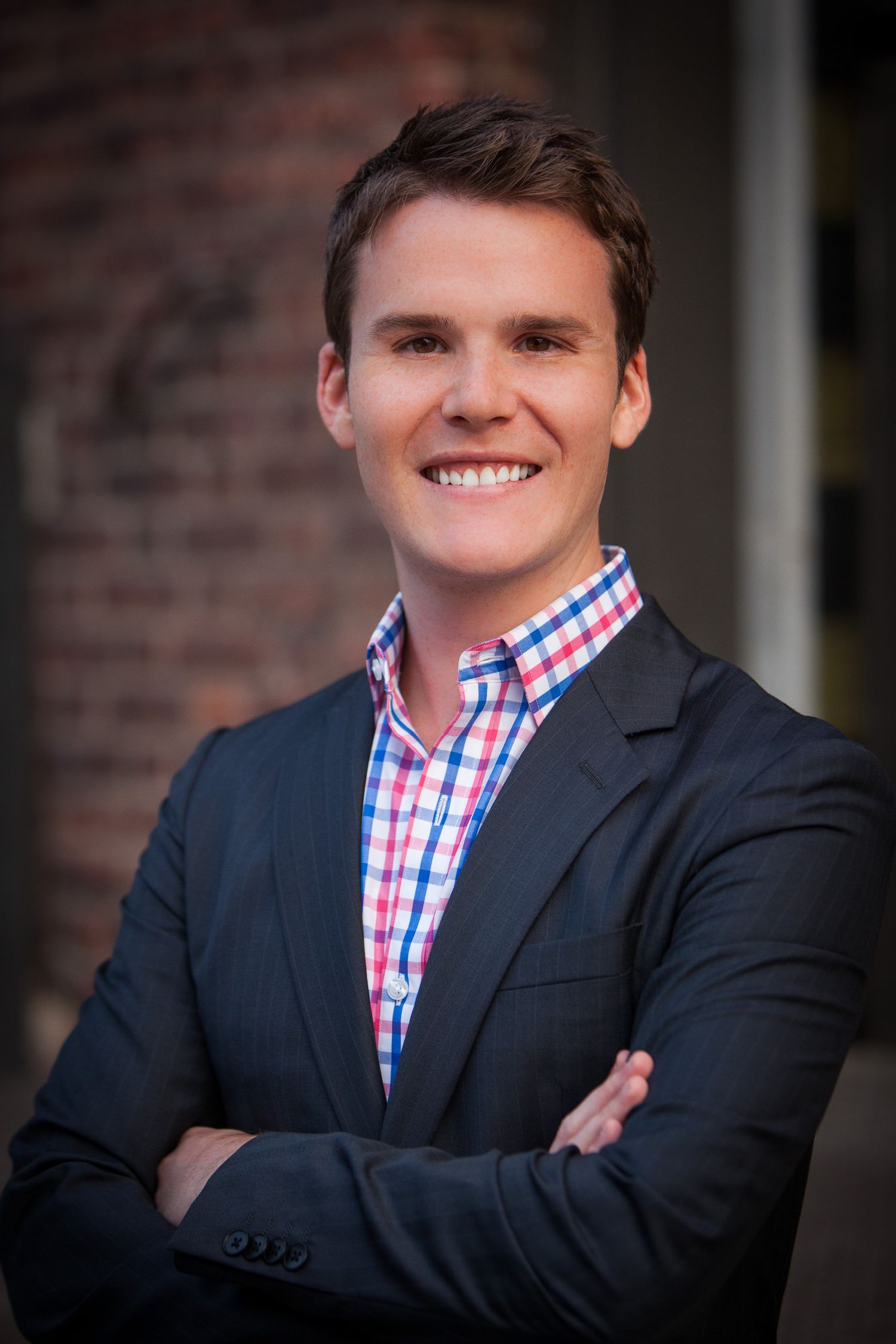 Young professional man in plaid shirt and navy blazer smiling with arms crossed against brick wall.
