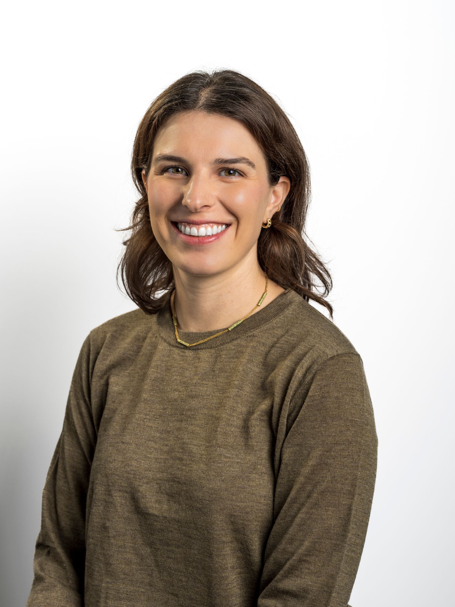 Woman with brown hair smiling, wearing an olive green long-sleeve top against a white background.