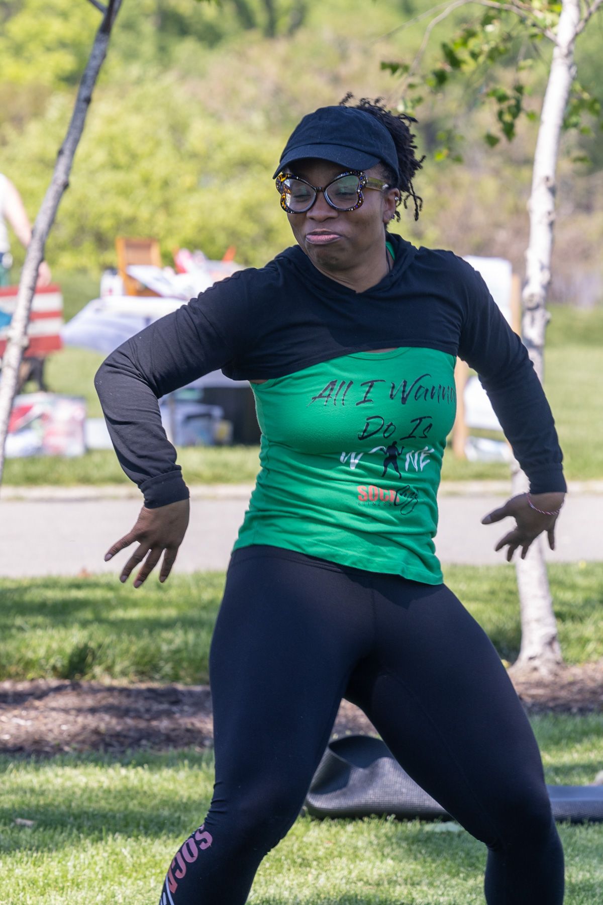 Woman in green shirt and black cap performs energetic dance moves outdoors in a park setting.