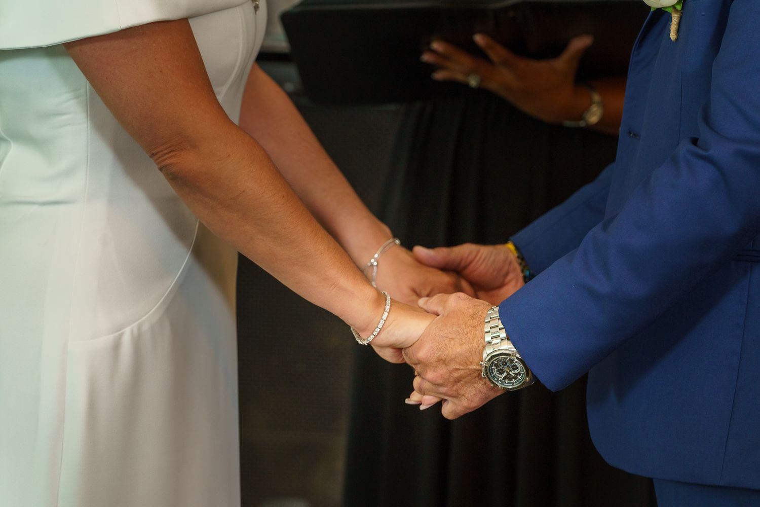 Couple holding hands at wedding ceremony, woman in white dress with bracelet, man in blue suit with watch.