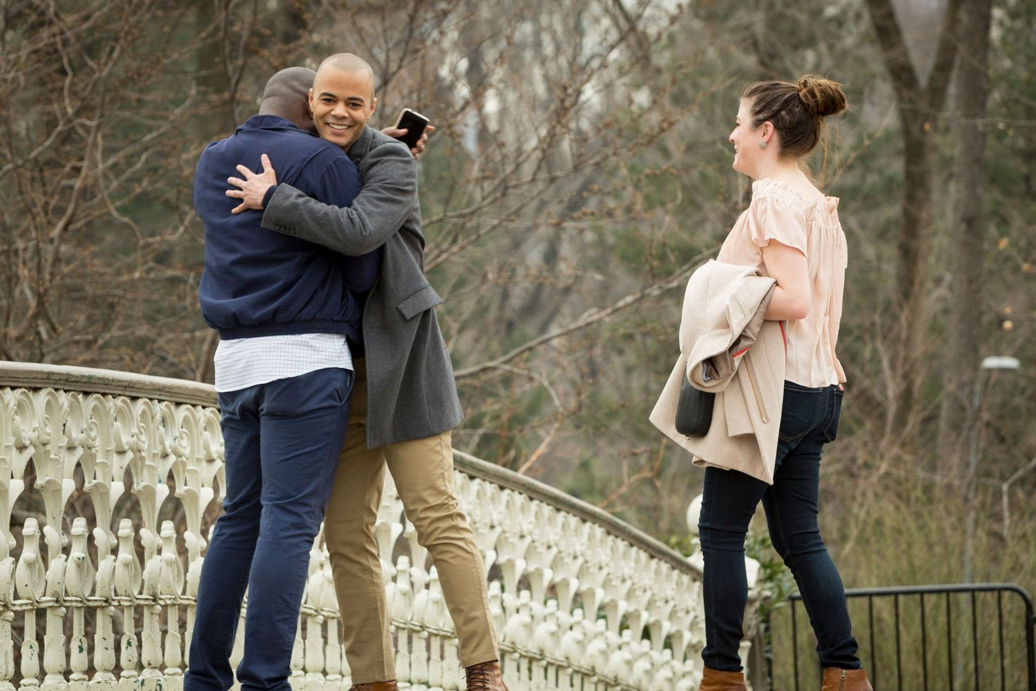 Two men hugging on a bridge in a park while a woman stands nearby holding a jacket outdoors.
