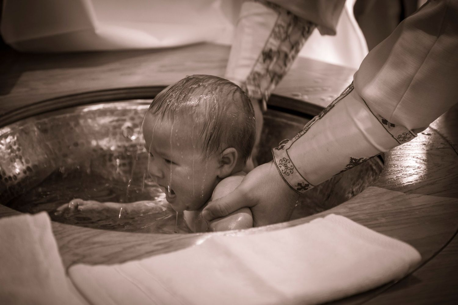 Infant being baptized in a ornate font, held by priest's hands during a Christian baptism ceremony.