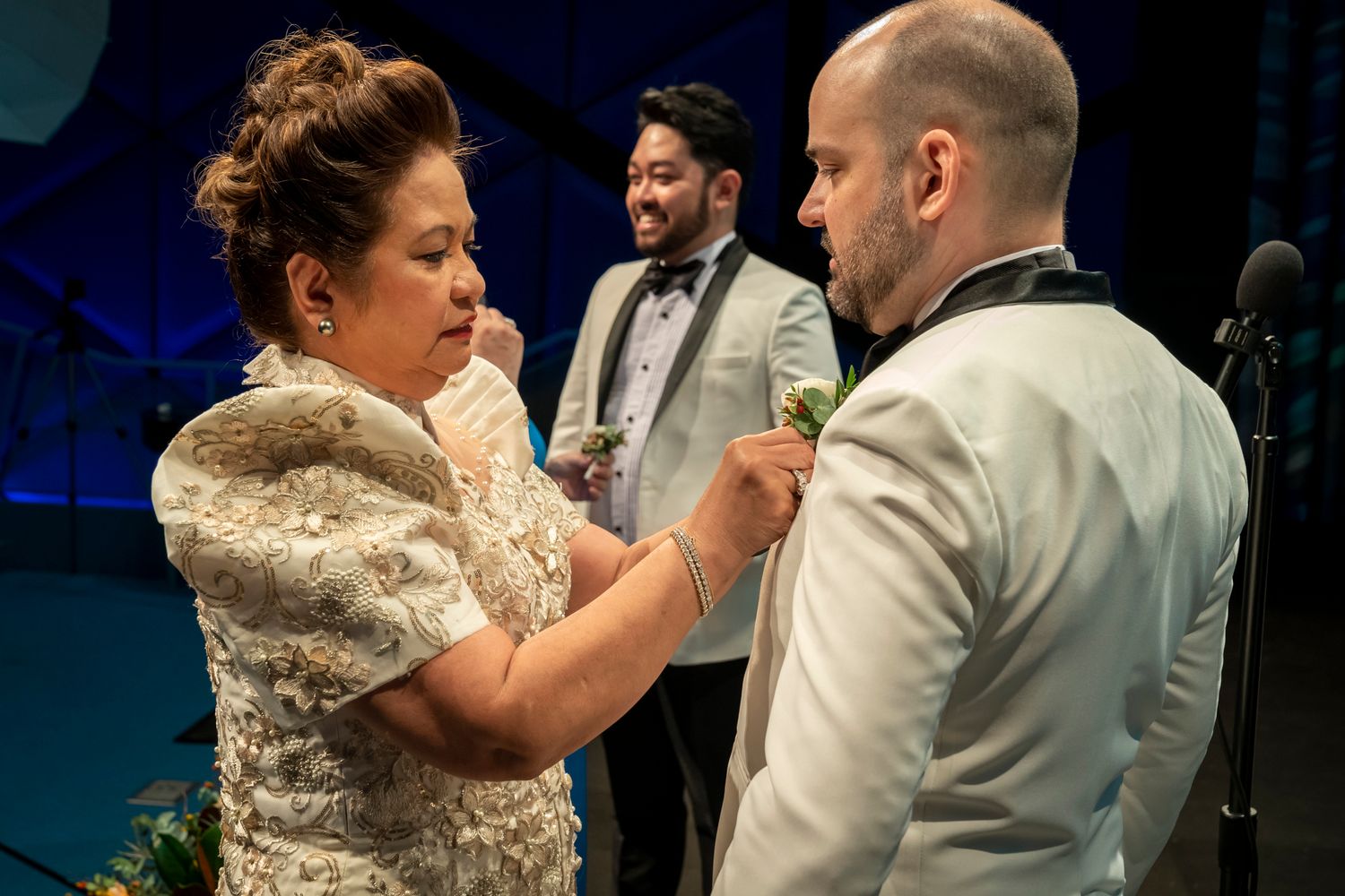 Woman pins boutonniere on groom's lapel at wedding ceremony while officiant stands in background.