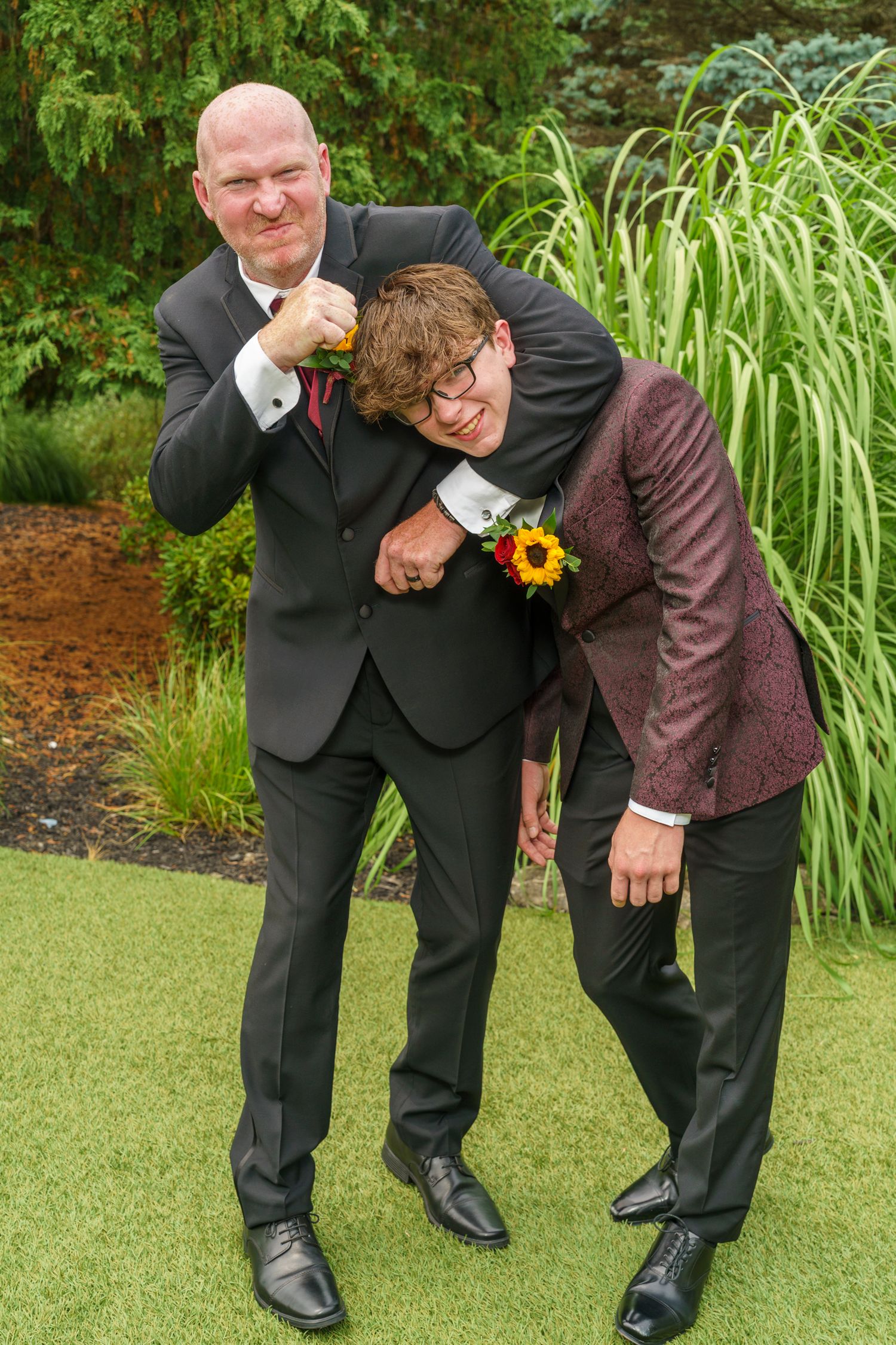 Two men in formal suits playfully pose outdoors at a wedding, one in a headlock, both wearing boutonnieres.
