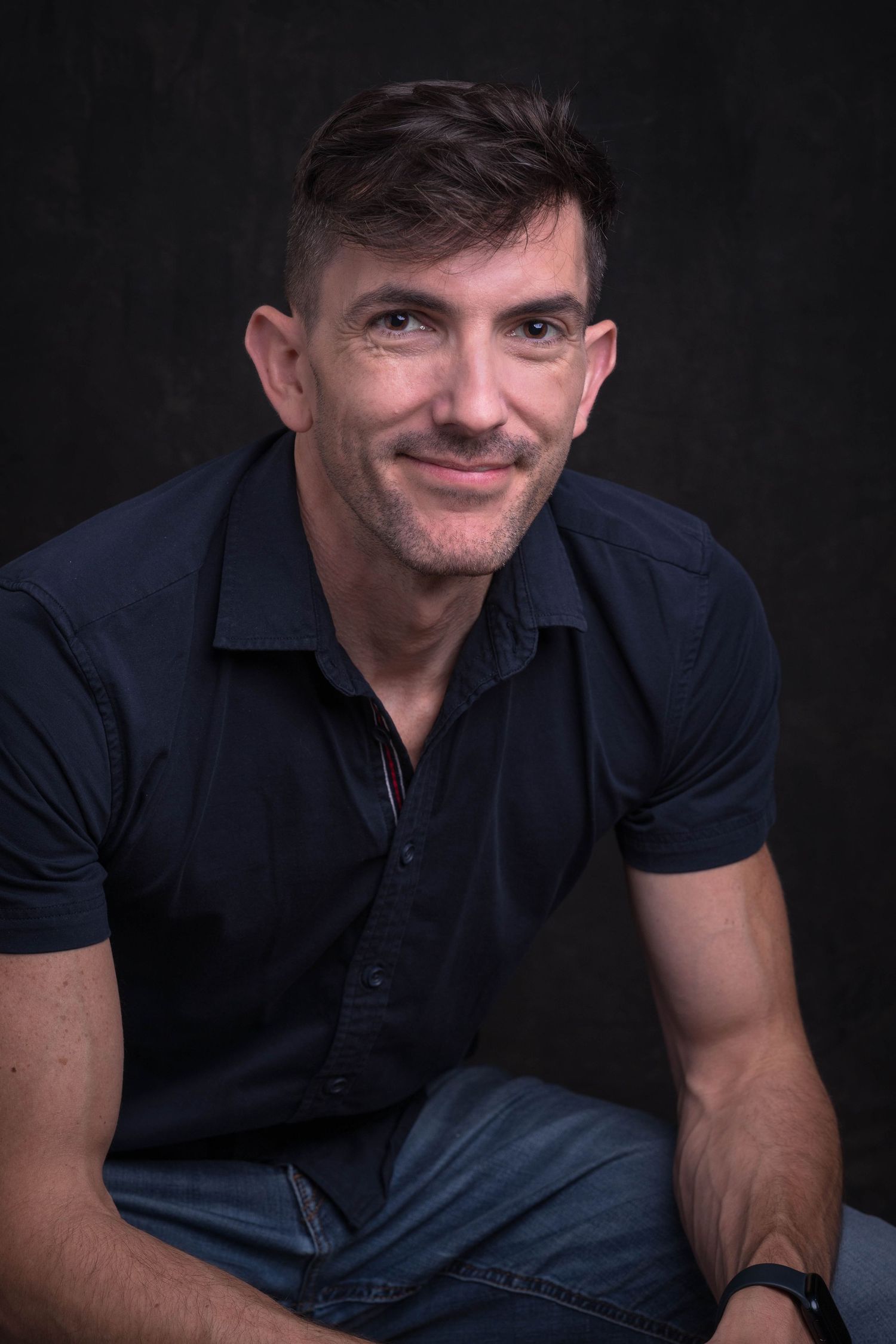 Middle-aged man in navy polo shirt smiling casually against dark background.