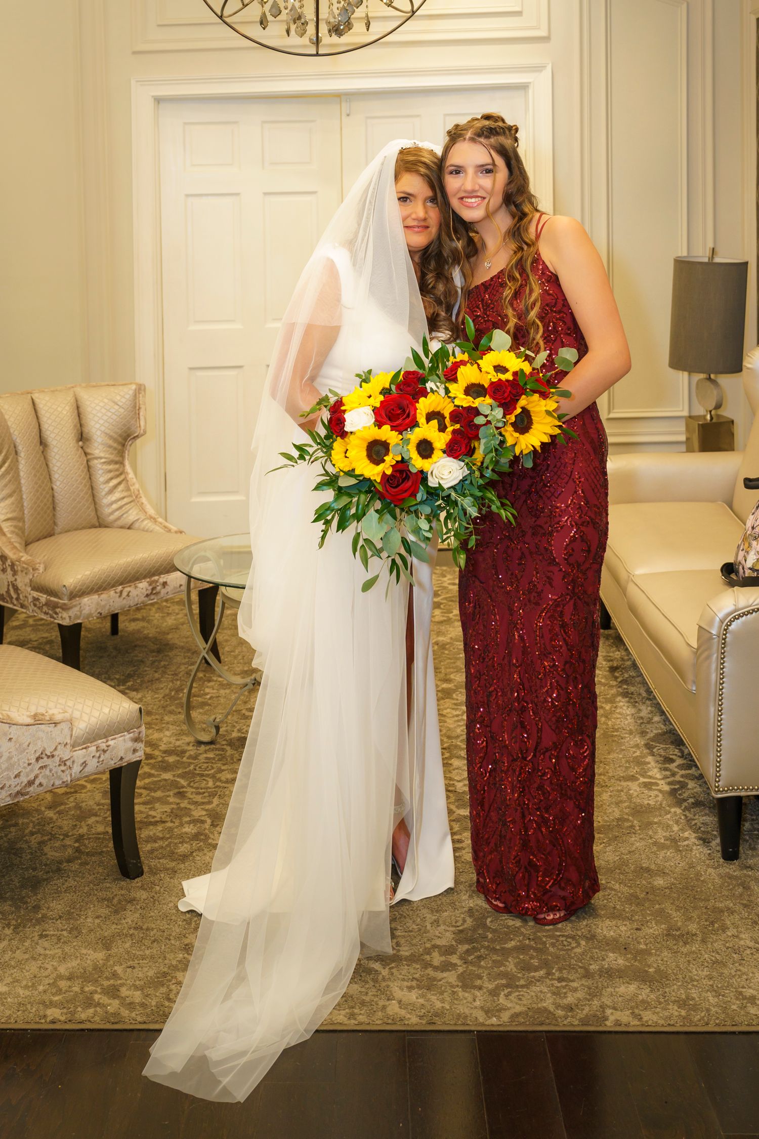 Bride in white gown with veil and bridesmaid in red dress holding sunflower and rose bouquet indoors.