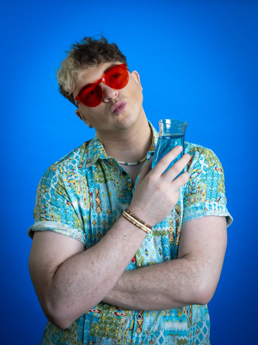 Young man in red sunglasses and colorful tropical shirt poses with blue vase against bright blue background.