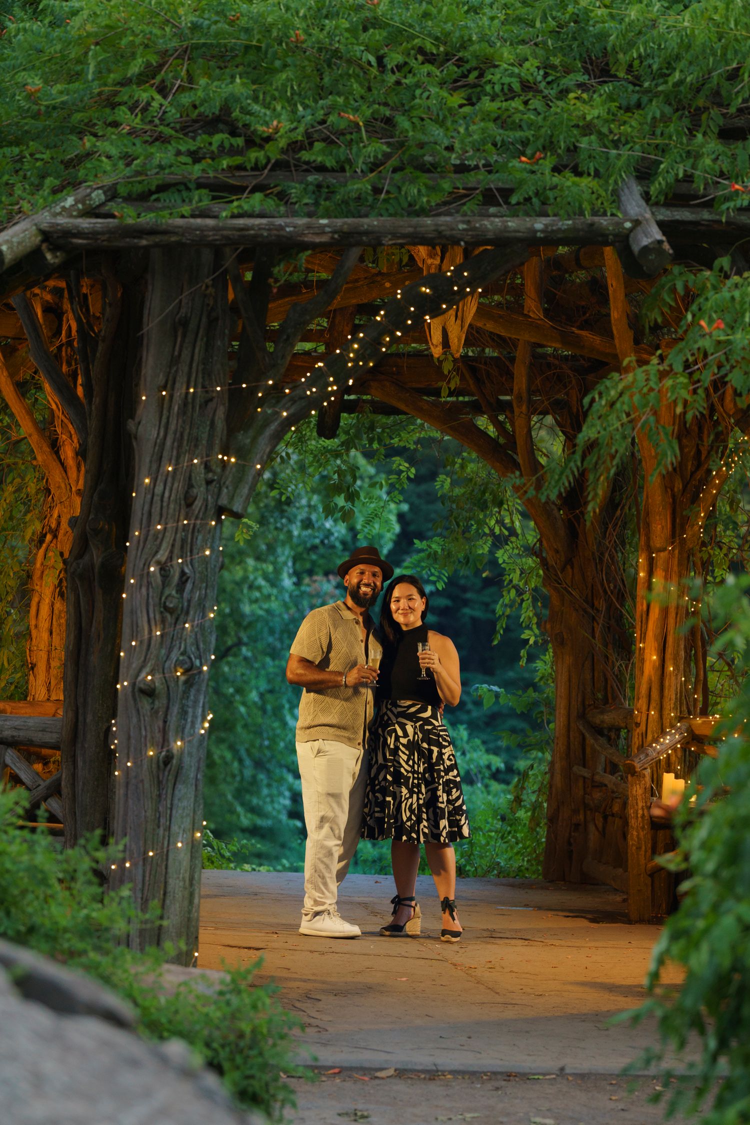 Couple smiling under a rustic wooden arbor with string lights at dusk in a lush garden setting.