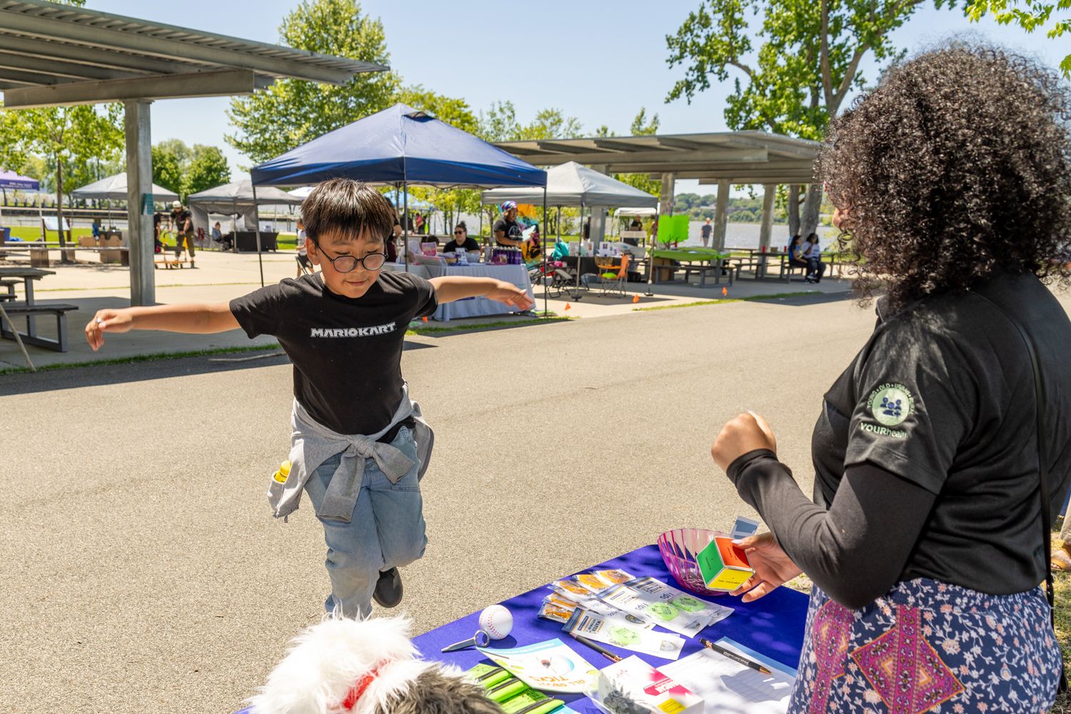 A child joyfully jumps at an outdoor community event booth while an adult volunteer smiles nearby.