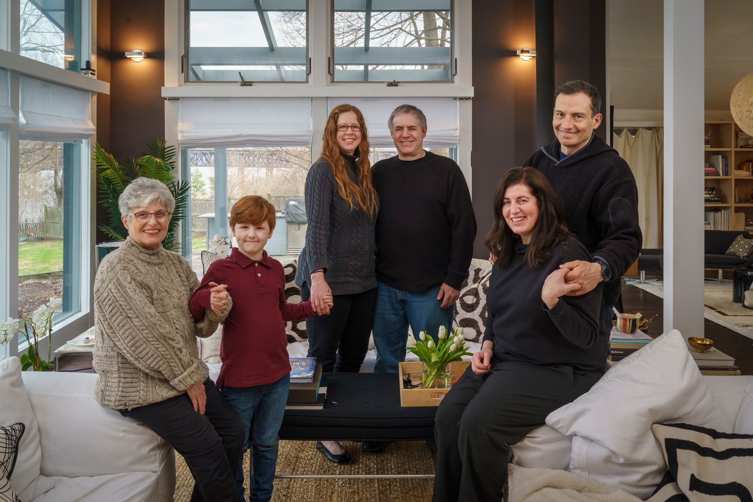 A multigenerational family of six smiling together in a modern living room with large windows and natural light.