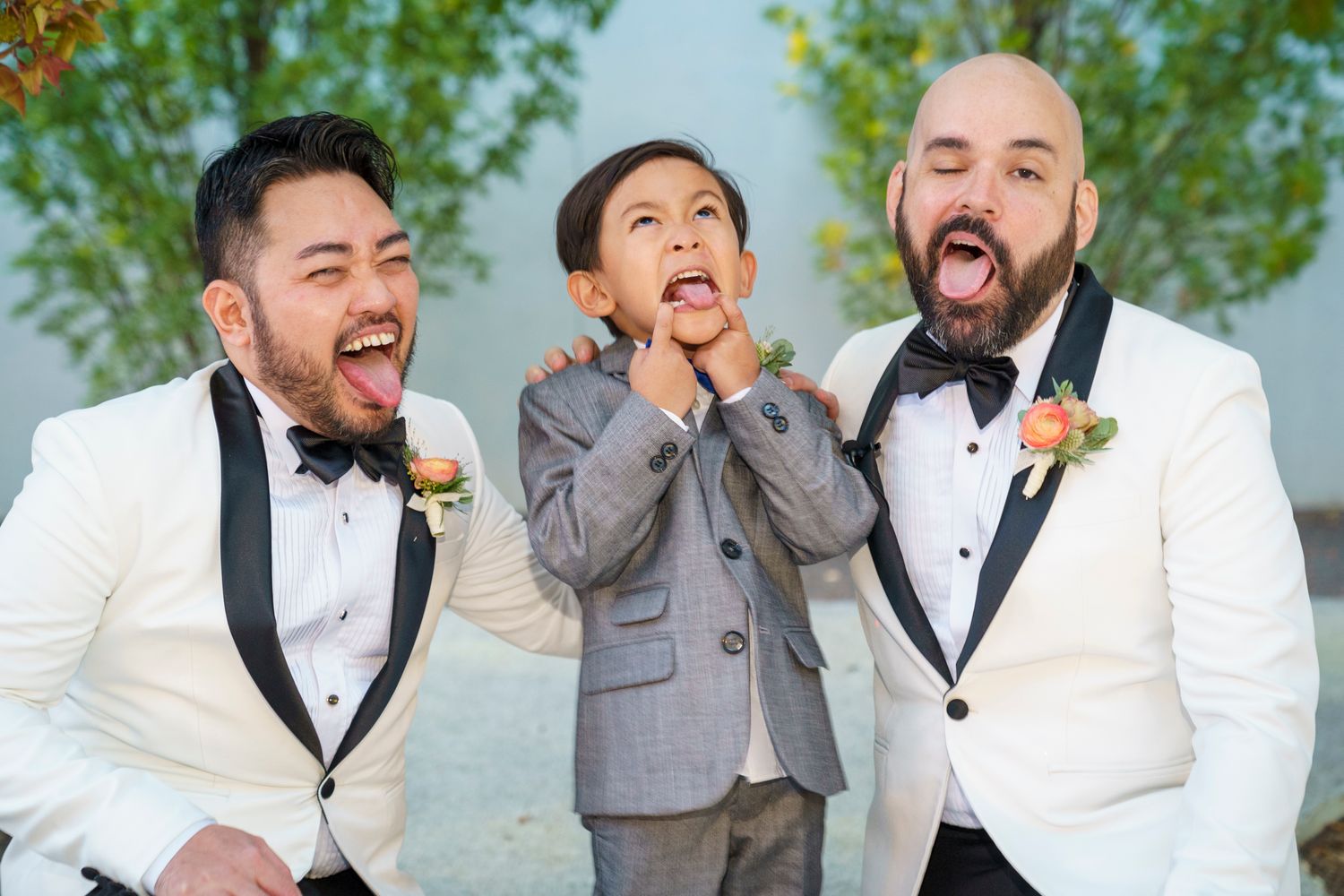 Two grooms in white tuxedos and a young boy in gray suit making funny faces at a wedding celebration.