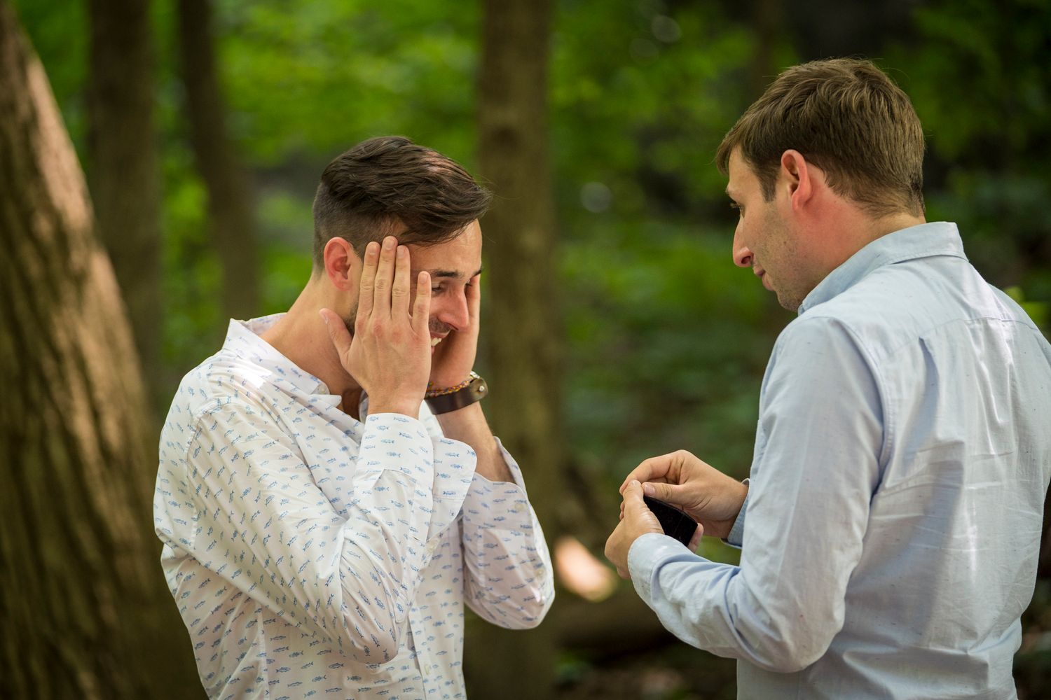 Emotional man covers face during outdoor marriage proposal while partner holds ring box in wooded setting.
