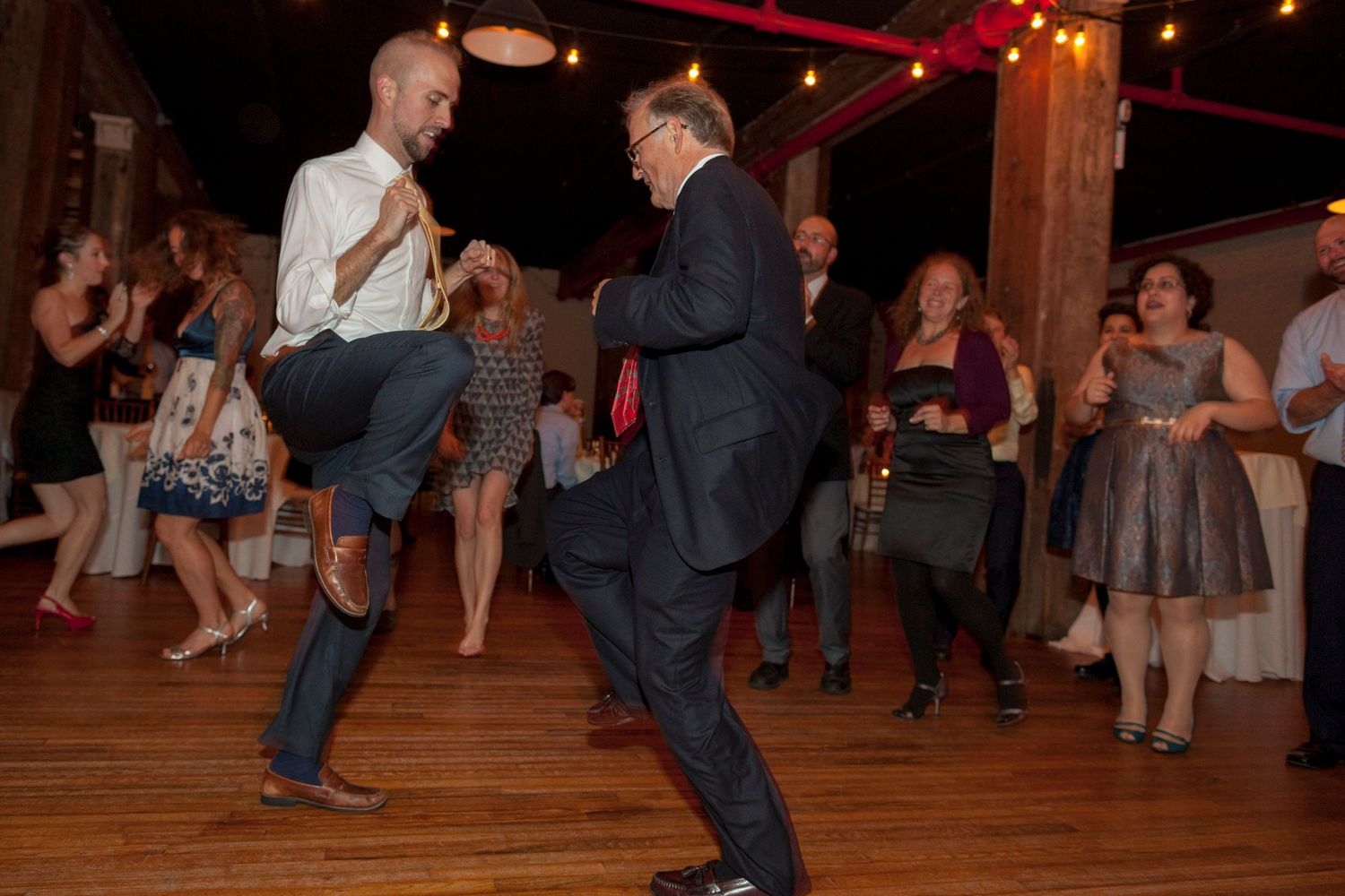 Two men energetically dancing at a rustic wedding reception while guests watch and celebrate.
