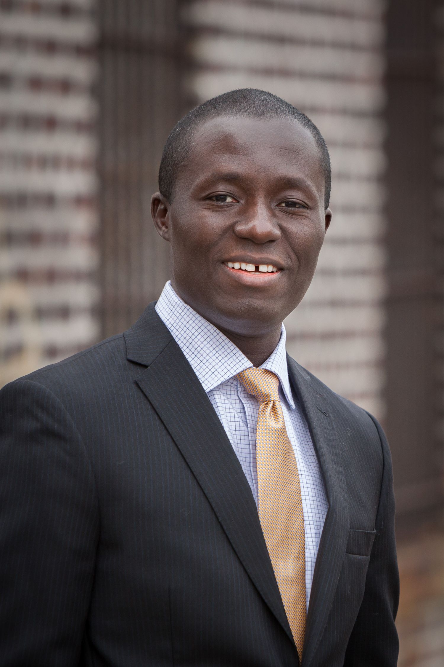 Young Black professional man in dark suit and gold tie smiling outdoors.