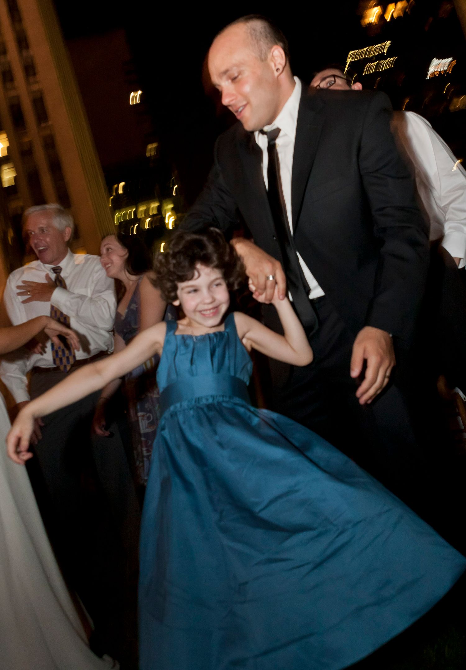 Man in black suit twirls young girl in teal dress on dance floor at evening wedding reception.