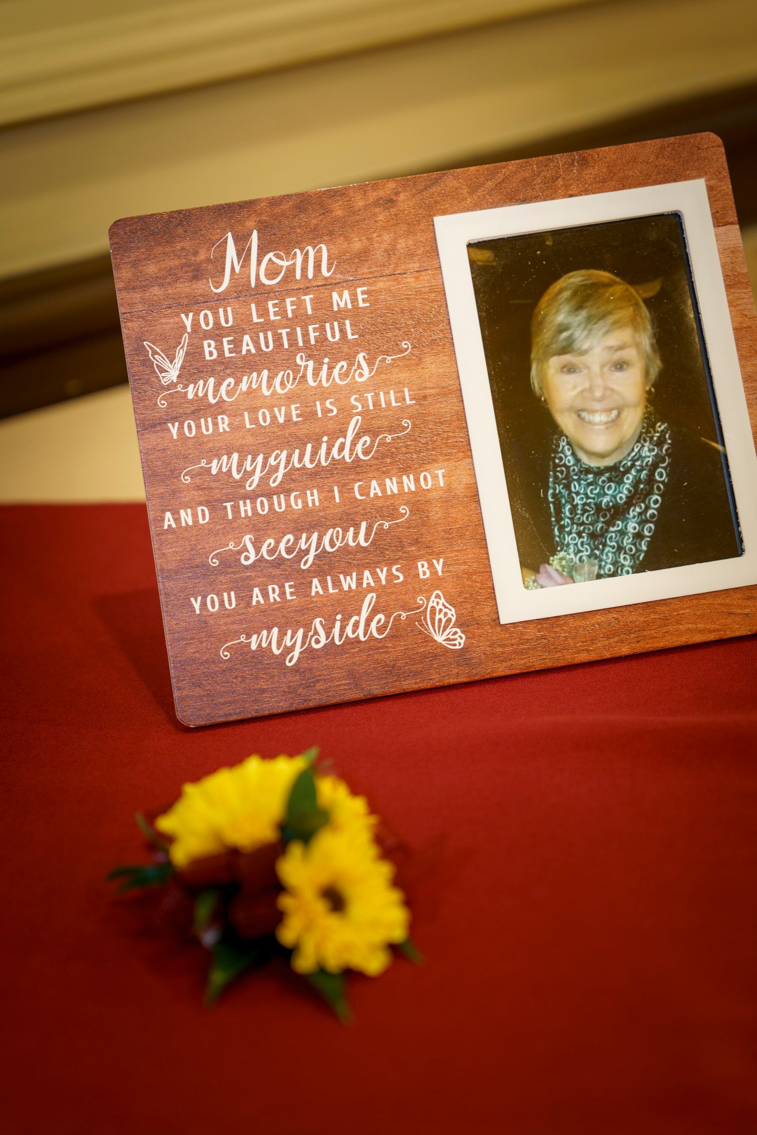 A memorial photo frame with a heartfelt Mom tribute message beside yellow flowers on a red tablecloth at a wedding reception.