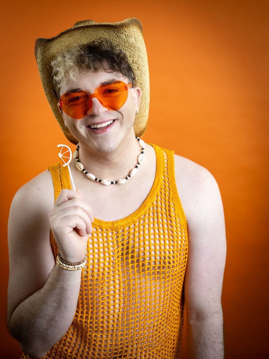Person wearing orange mesh top, cowboy hat, orange sunglasses, and bead necklace holding a lollipop on orange background.