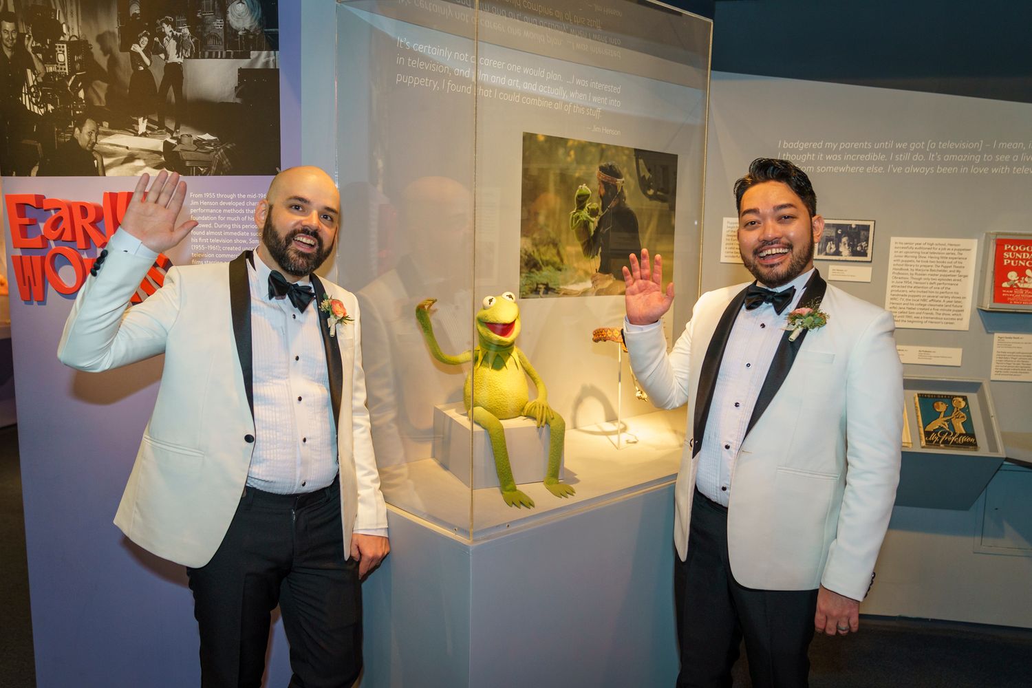Two men in white tuxedos wave beside a Kermit the Frog display case at a museum exhibit.