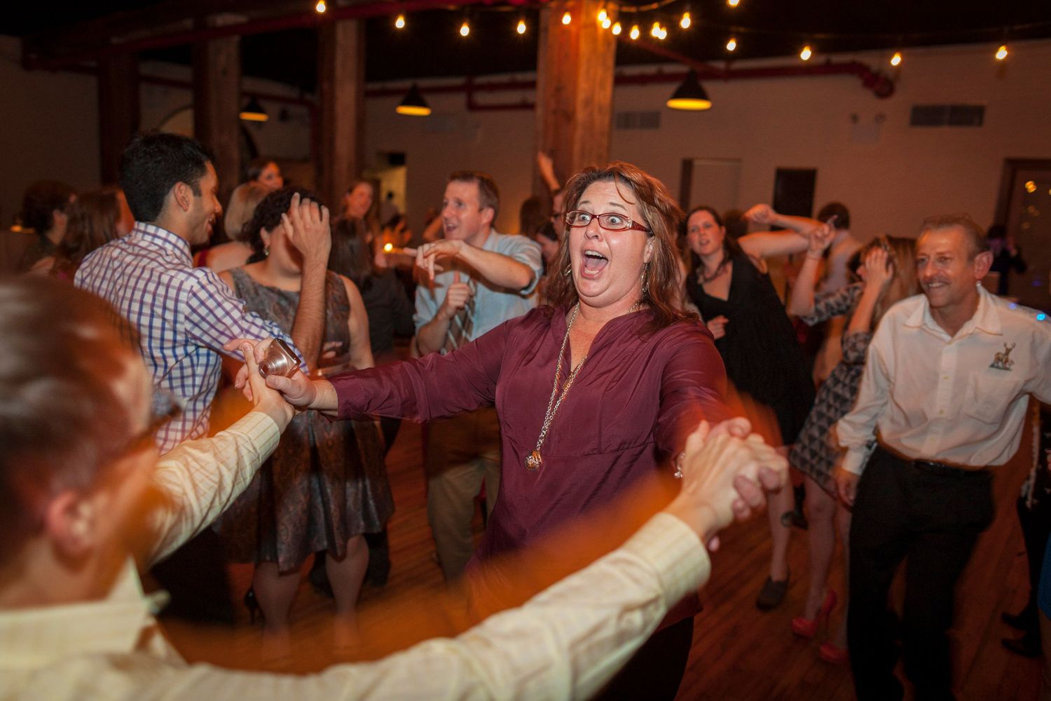 Guests dancing joyfully at a lively indoor wedding reception with warm string lights overhead.