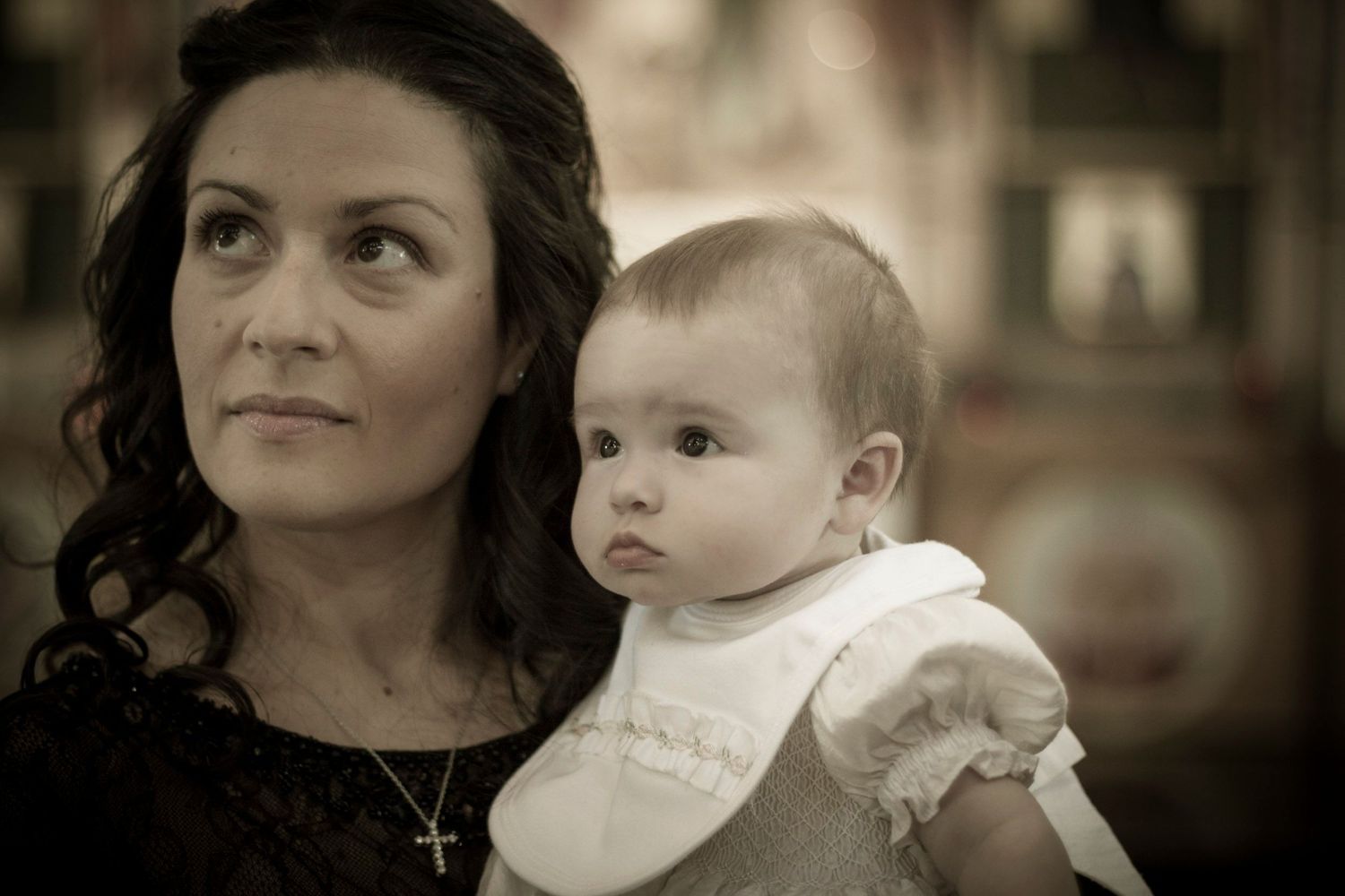 A woman wearing a gold cross necklace holds a baby dressed in white during what appears to be a baptism ceremony.