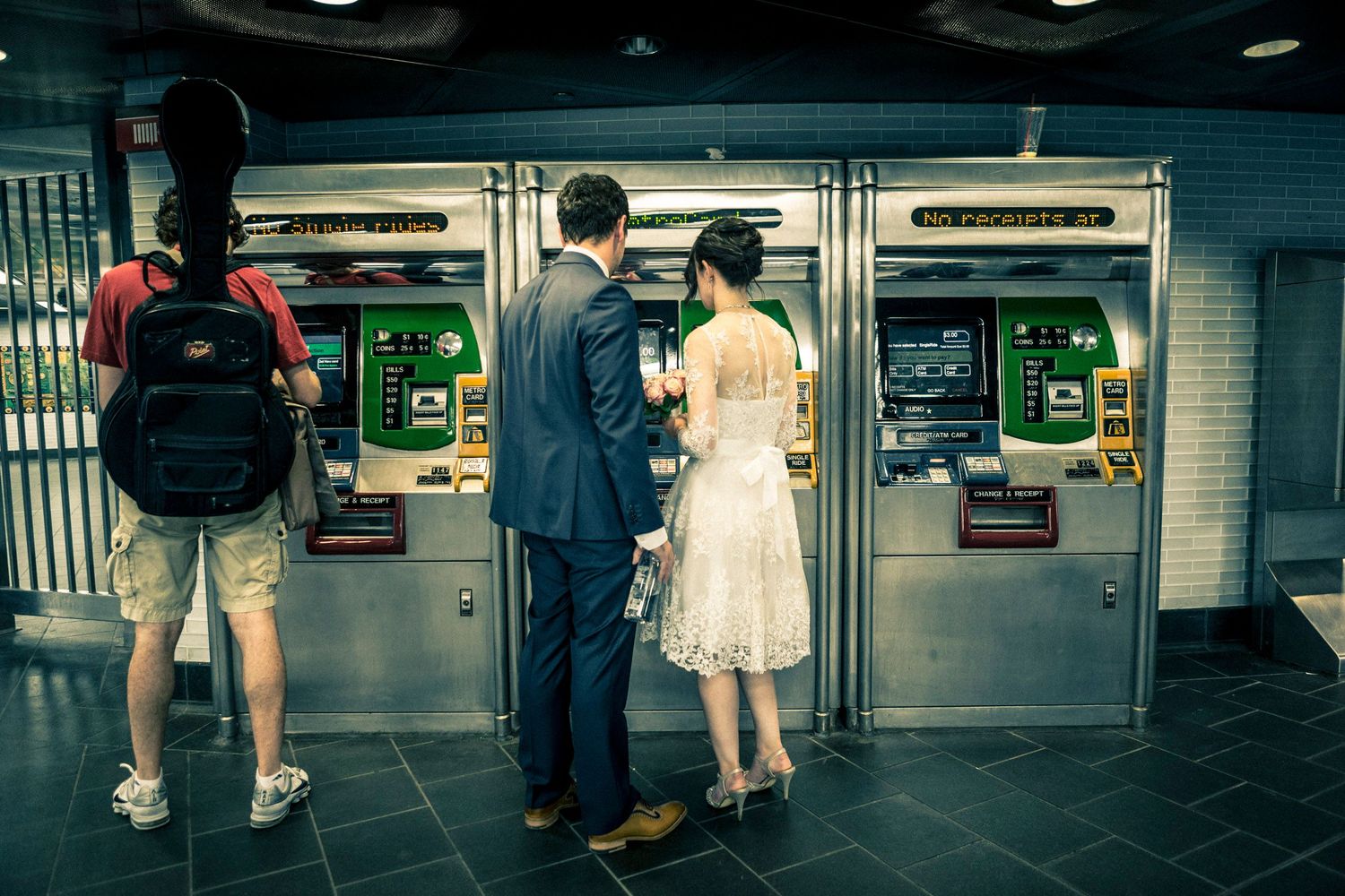 Couple in formal attire using a subway ticket ATM machine, with another traveler carrying a red backpack nearby.