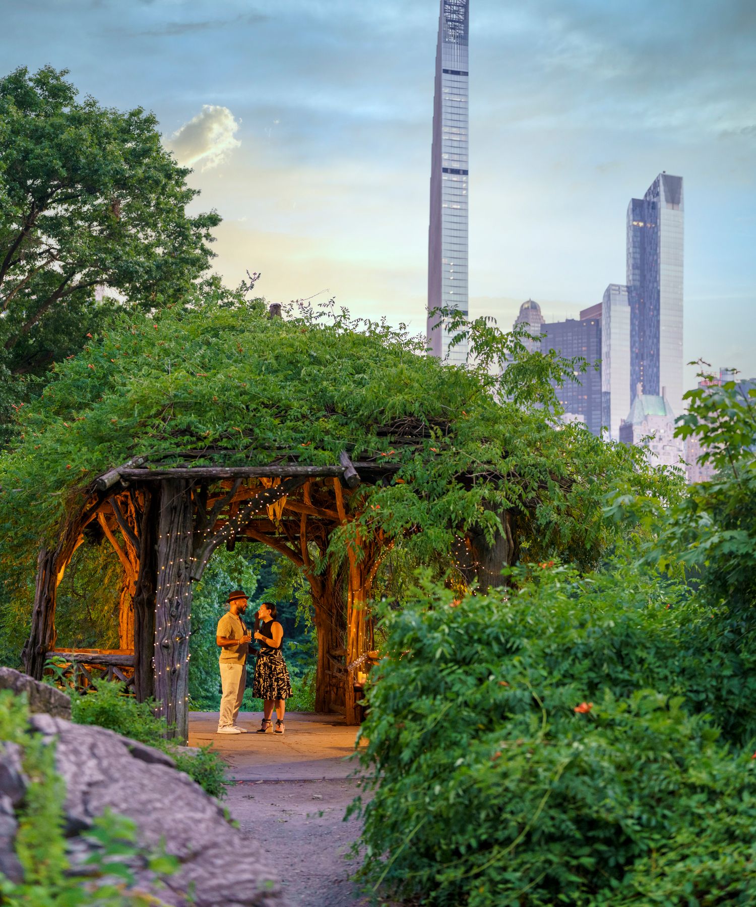 Couple embracing under ivy-covered archway in Central Park with NYC skyscrapers at dusk.