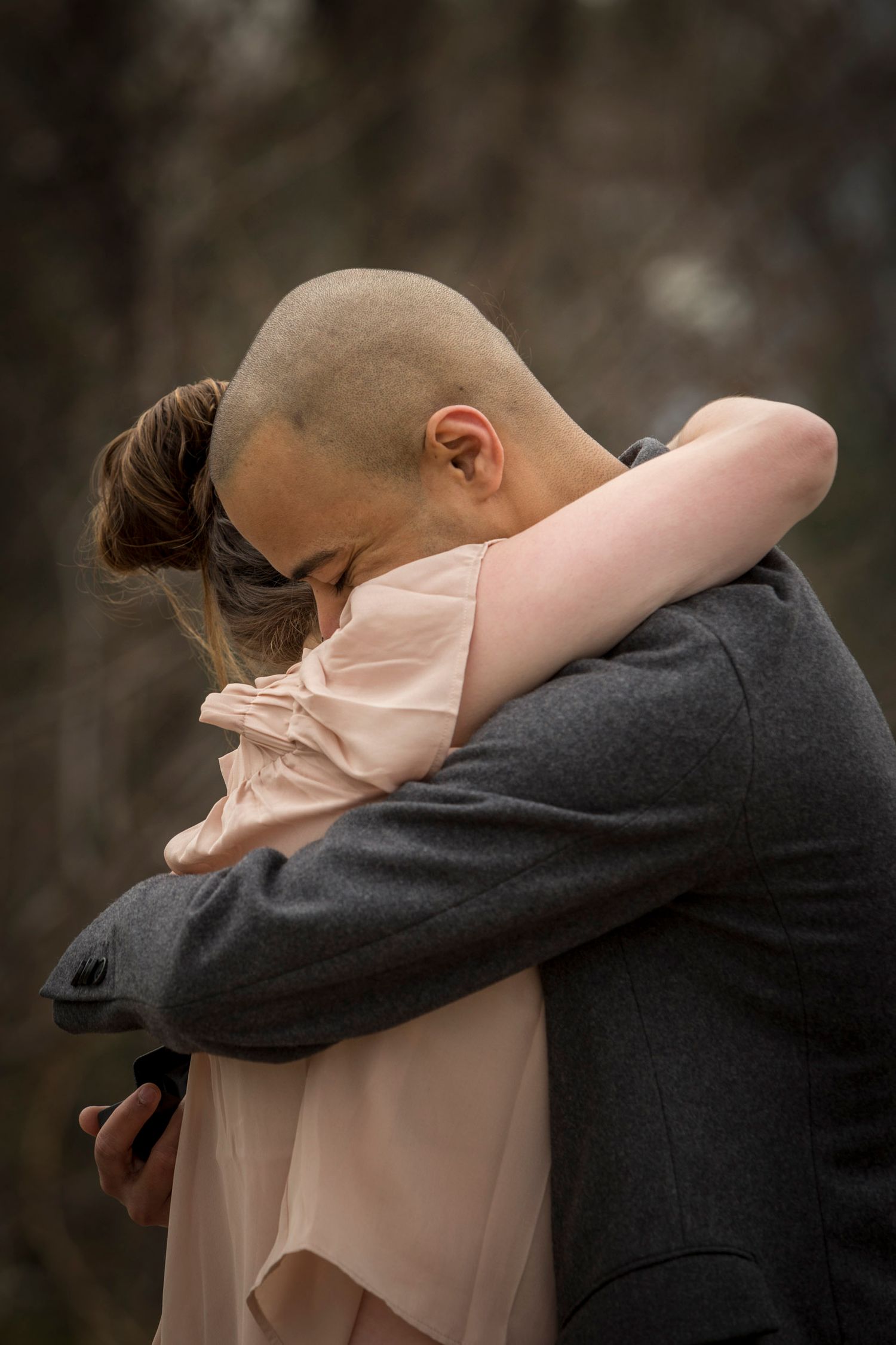 A bald man and a woman in a pink dress share a tender embrace outdoors in a blurred forest setting.