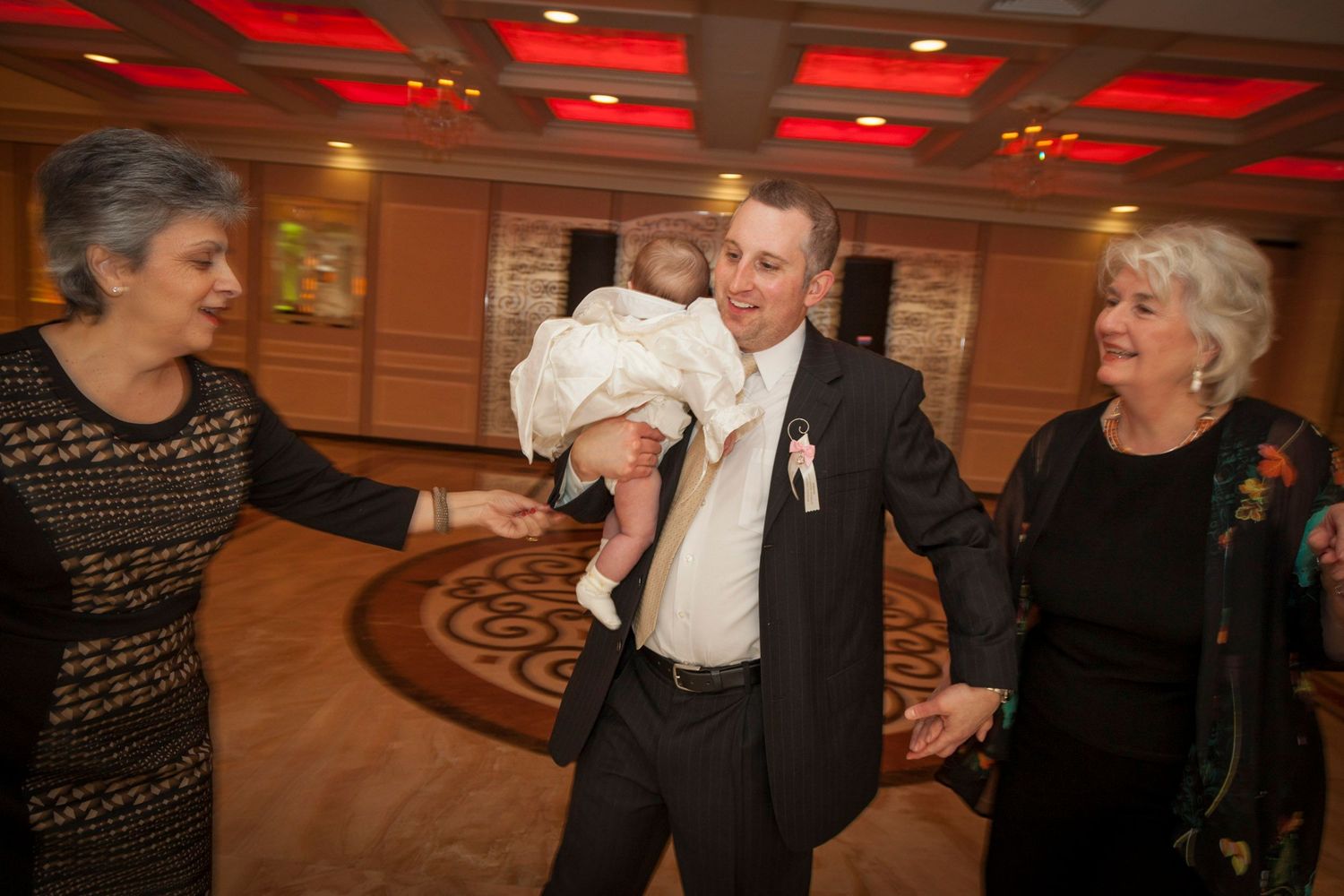 Man holding baby dances with two women in elegant ballroom at formal event.