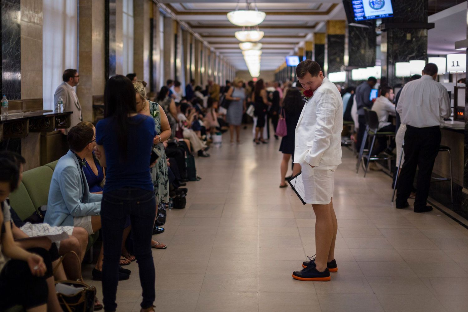 Busy government office waiting room with people seated and standing in a grand hall with ornate ceiling lights.