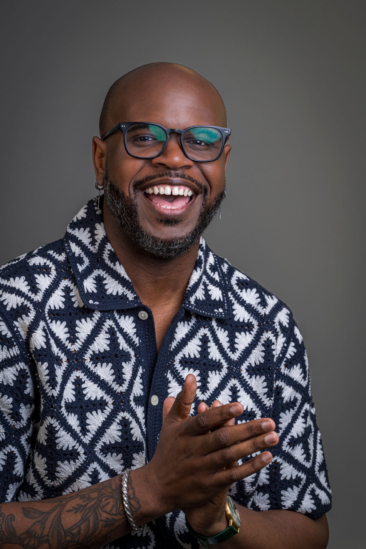 Smiling bald man with glasses and beard wearing a patterned blue shirt, clapping hands, with tattoos and bracelet.