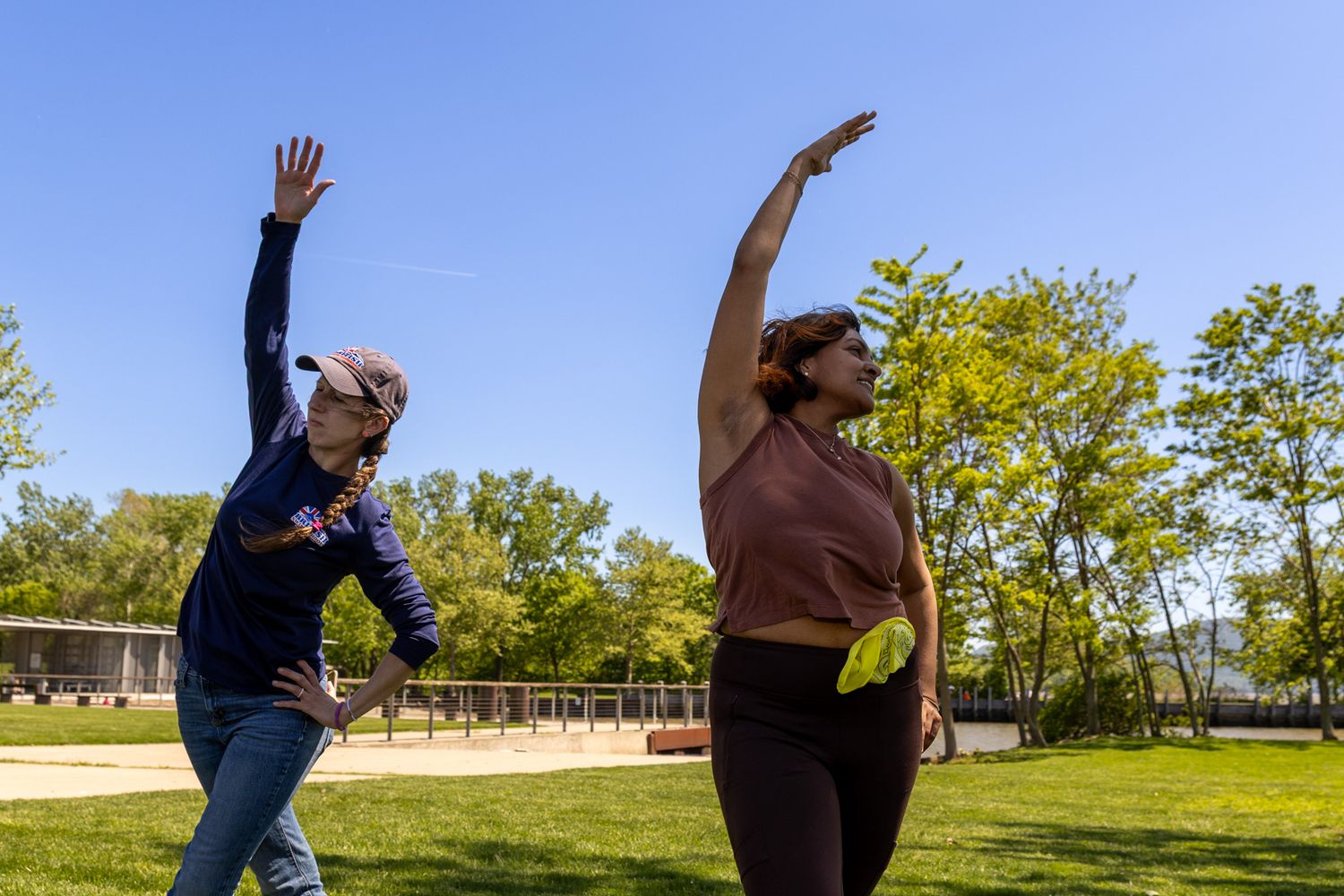 Two women stretching with arms raised during an outdoor fitness class in a sunny park.