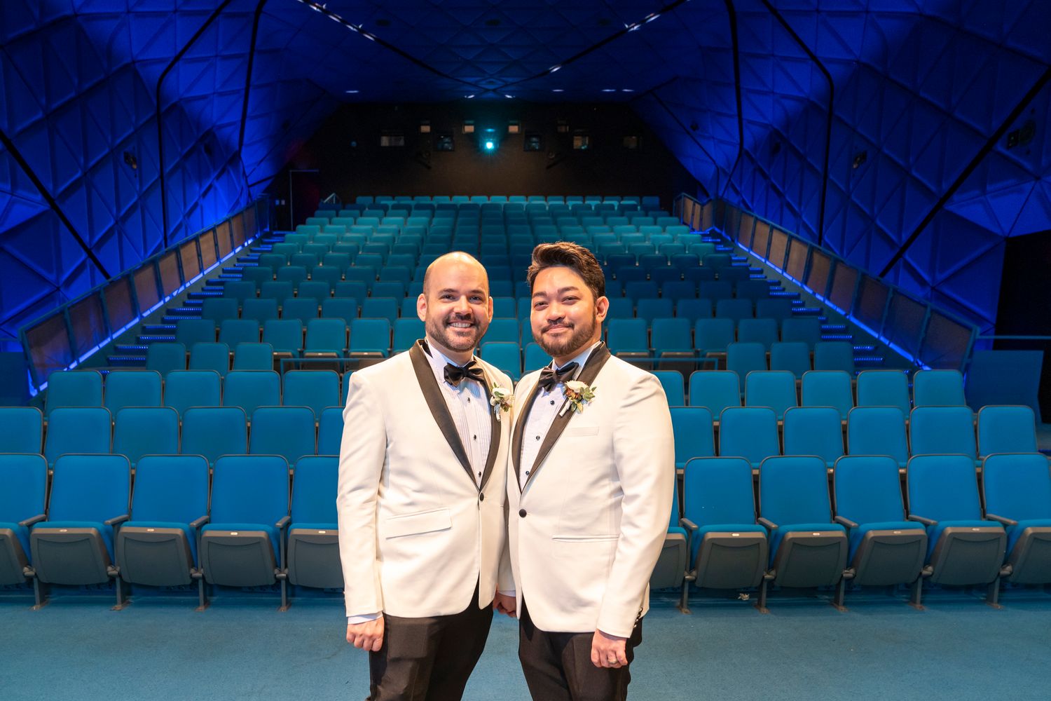 Two men in white tuxedos pose in a blue-lit theater with rows of empty blue seats behind them.