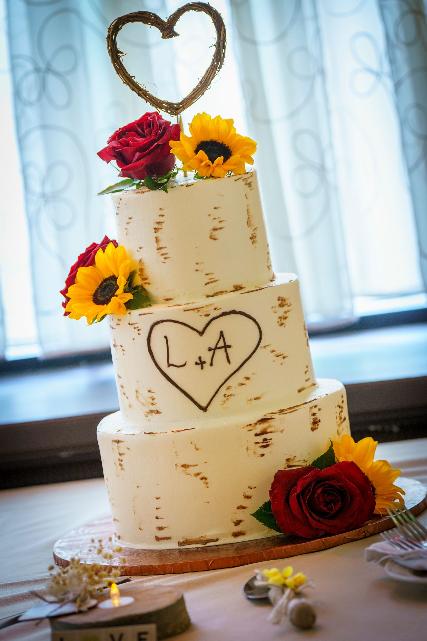 Three-tier rustic wedding cake with sunflowers and red roses, featuring carved L+A initials and a twig heart topper.