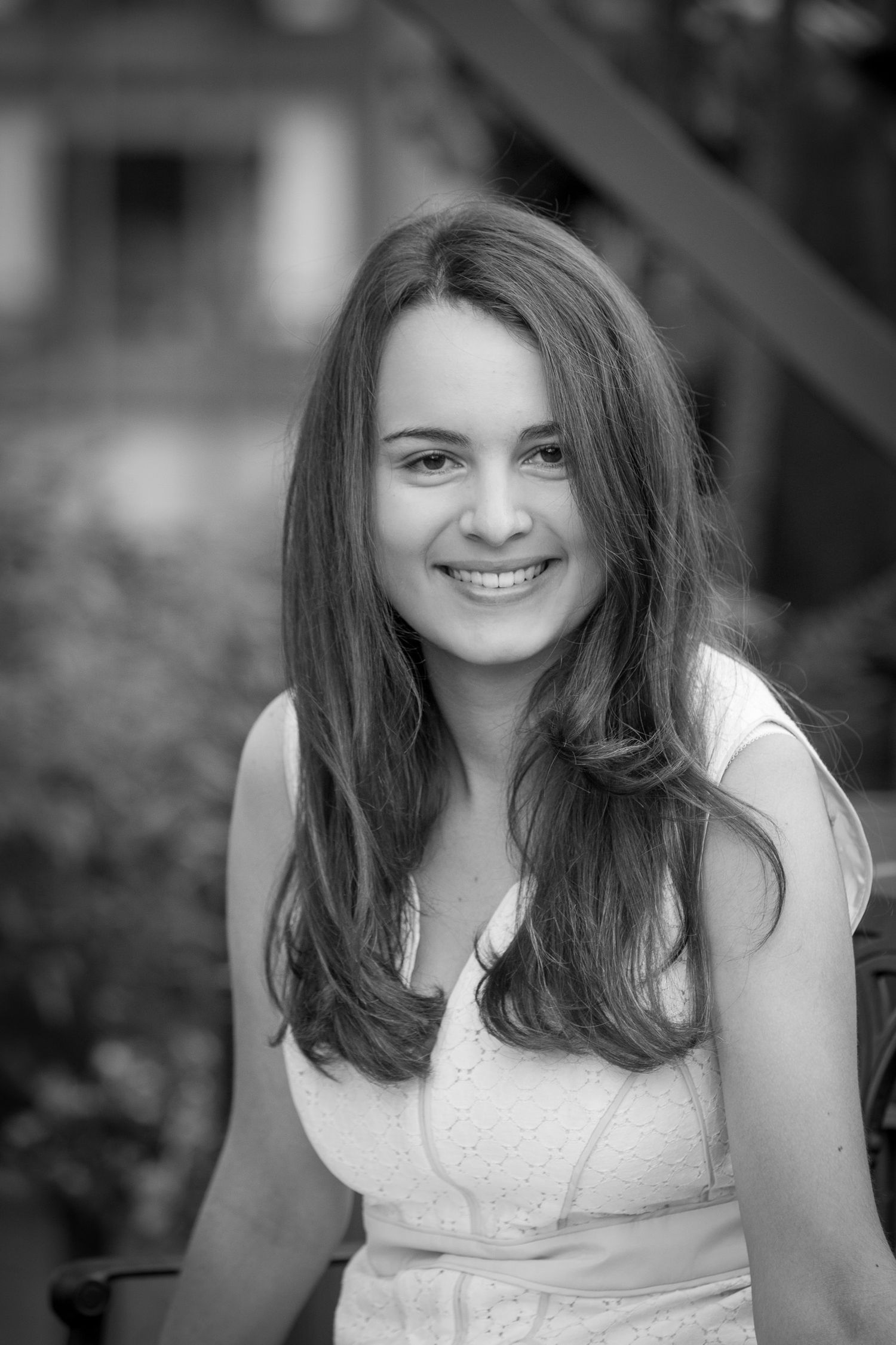 Black and white portrait of a smiling young woman with long hair wearing a white dress outdoors.