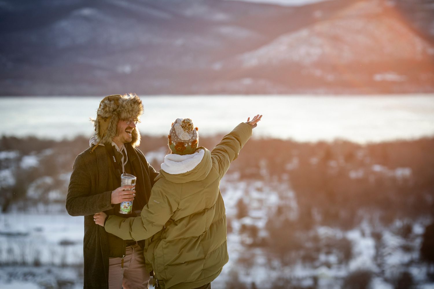 Two people enjoying a snowy winter landscape at sunset, one pointing toward a frozen lake and mountains.