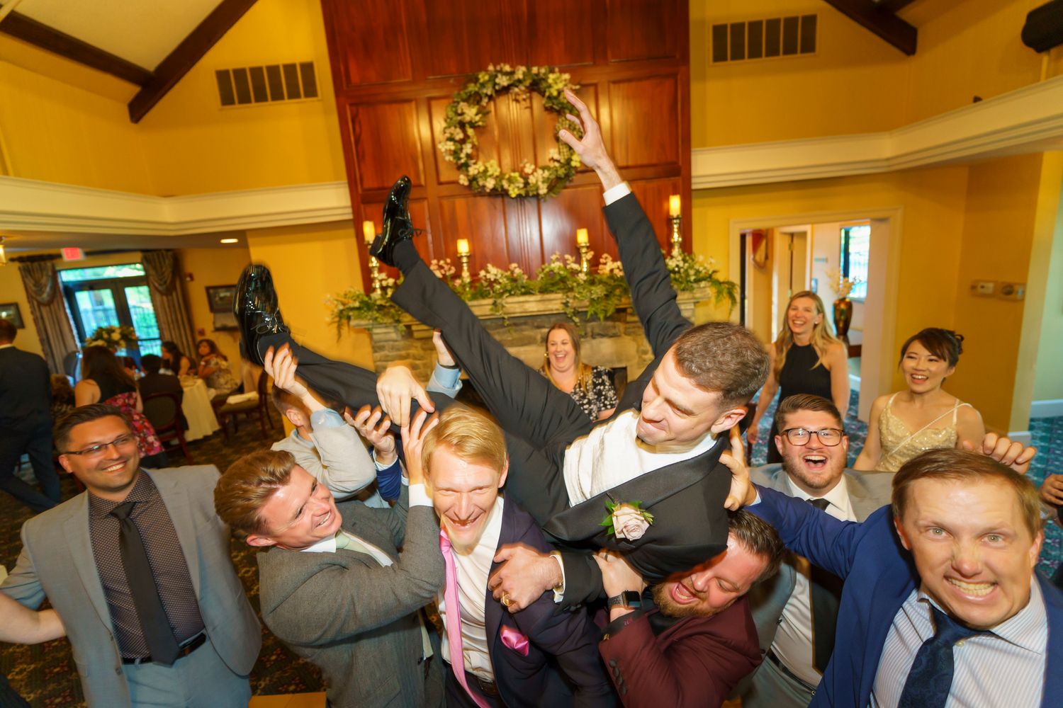 Excited groom lifted by groomsmen in suits celebrating at a lively indoor wedding reception party.