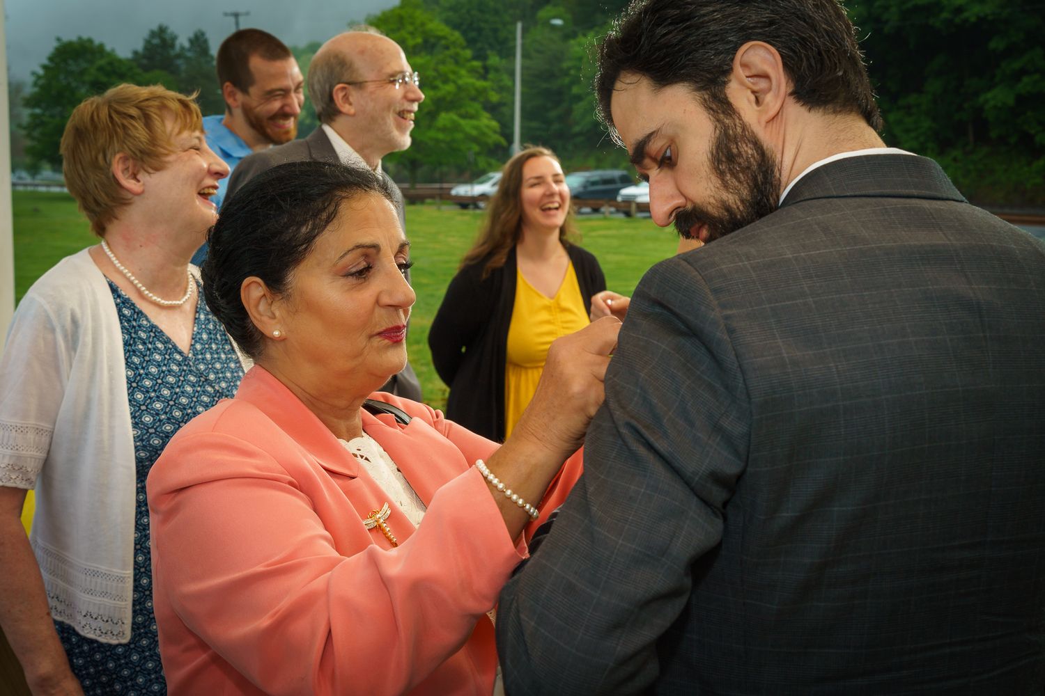 Woman in pink jacket pins boutonniere on man in suit outdoors, surrounded by smiling guests at a celebration.