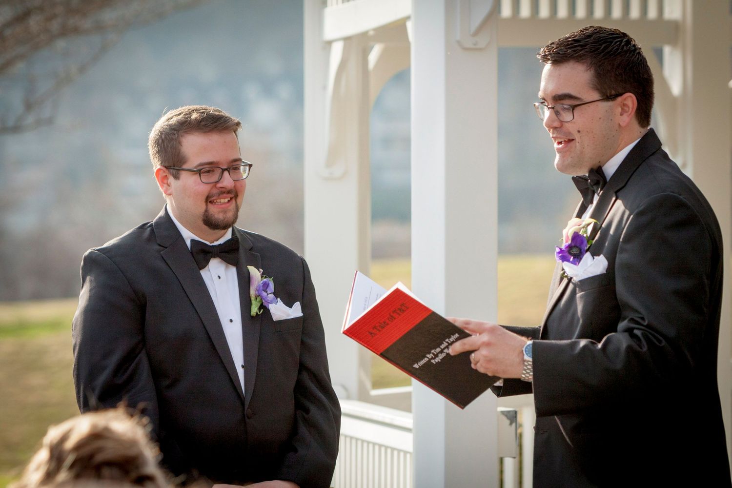 Two grooms in tuxedos exchange vows at an outdoor wedding ceremony beneath a white gazebo.