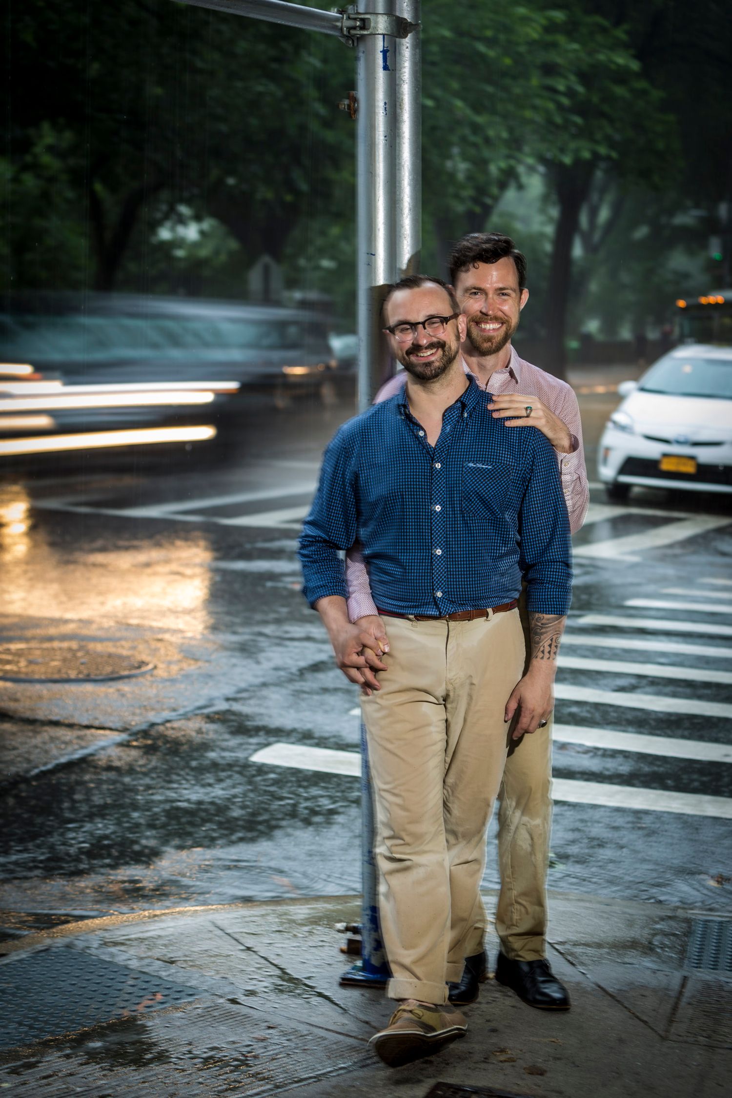 Two smiling men stand together on a rainy city street corner, one with a tattoo, both wearing casual button-up shirts.