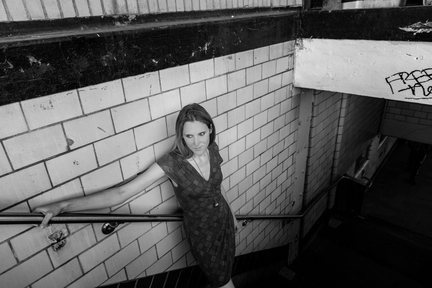 Black and white photo of a woman in a dress leaning on a railing in an urban subway stairwell.