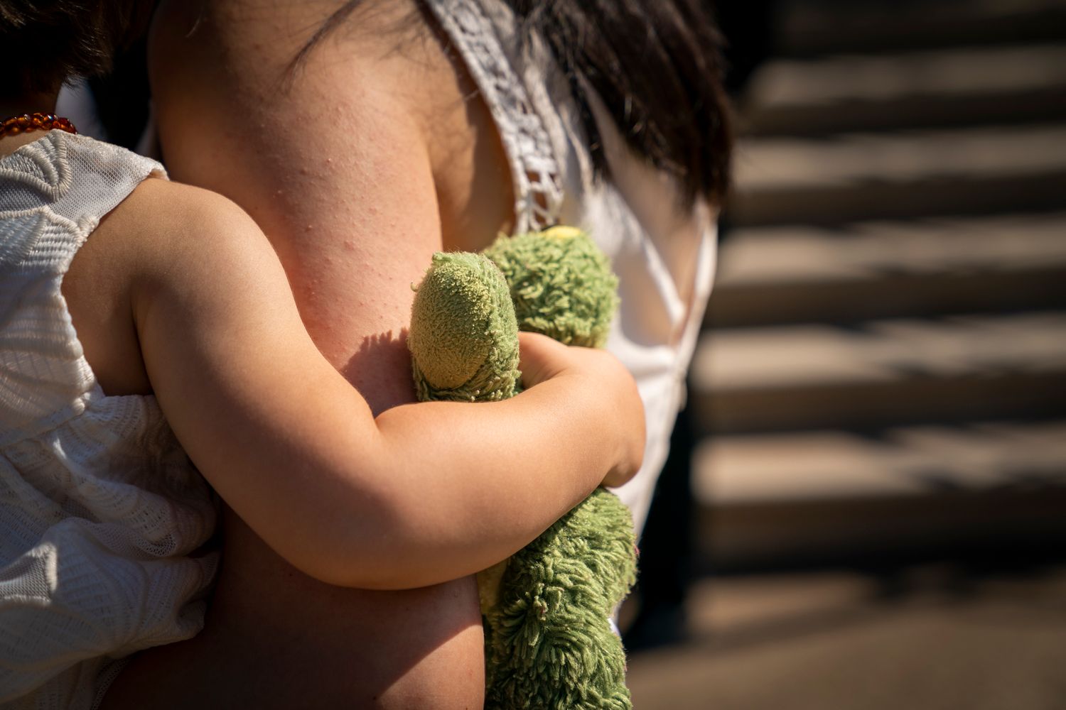 Child hugging a green stuffed teddy bear while being held by an adult outdoors on sunny steps.