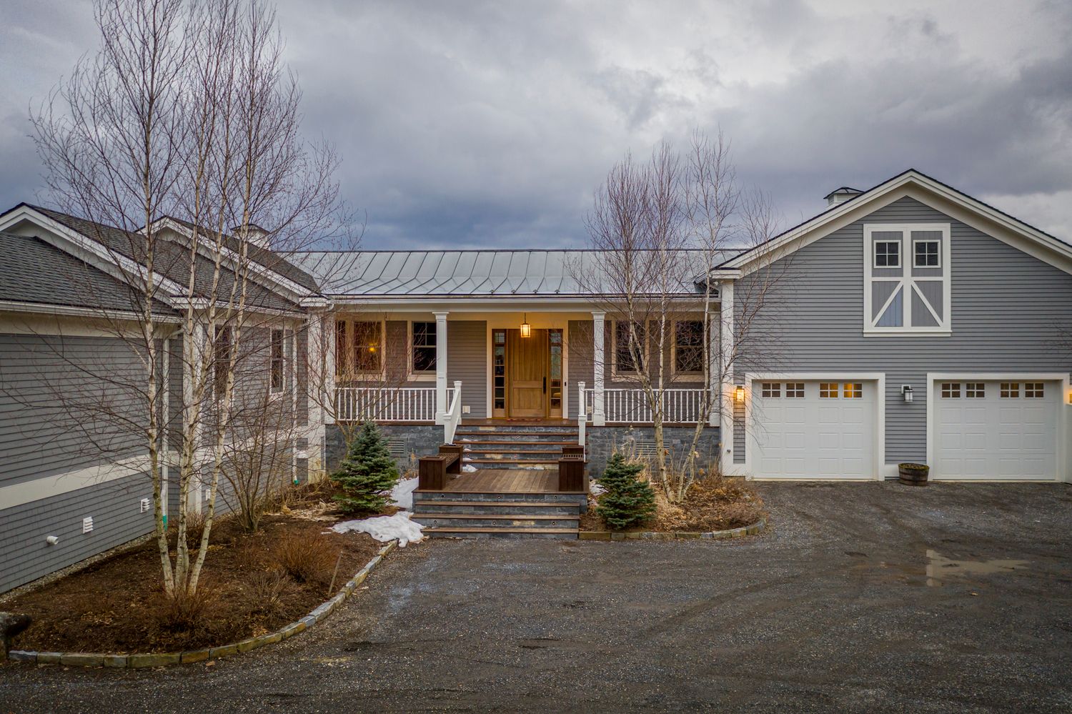 Gray farmhouse with covered porch, white-trimmed garage, metal roof, and bare birch trees on a cloudy winter day.