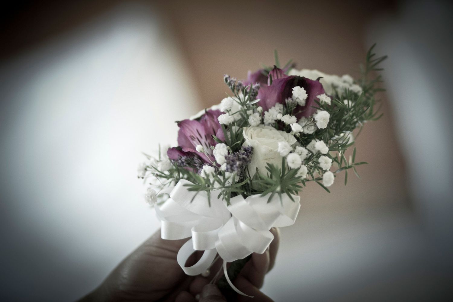 A hand holding a small floral corsage with purple flowers and white ribbon.