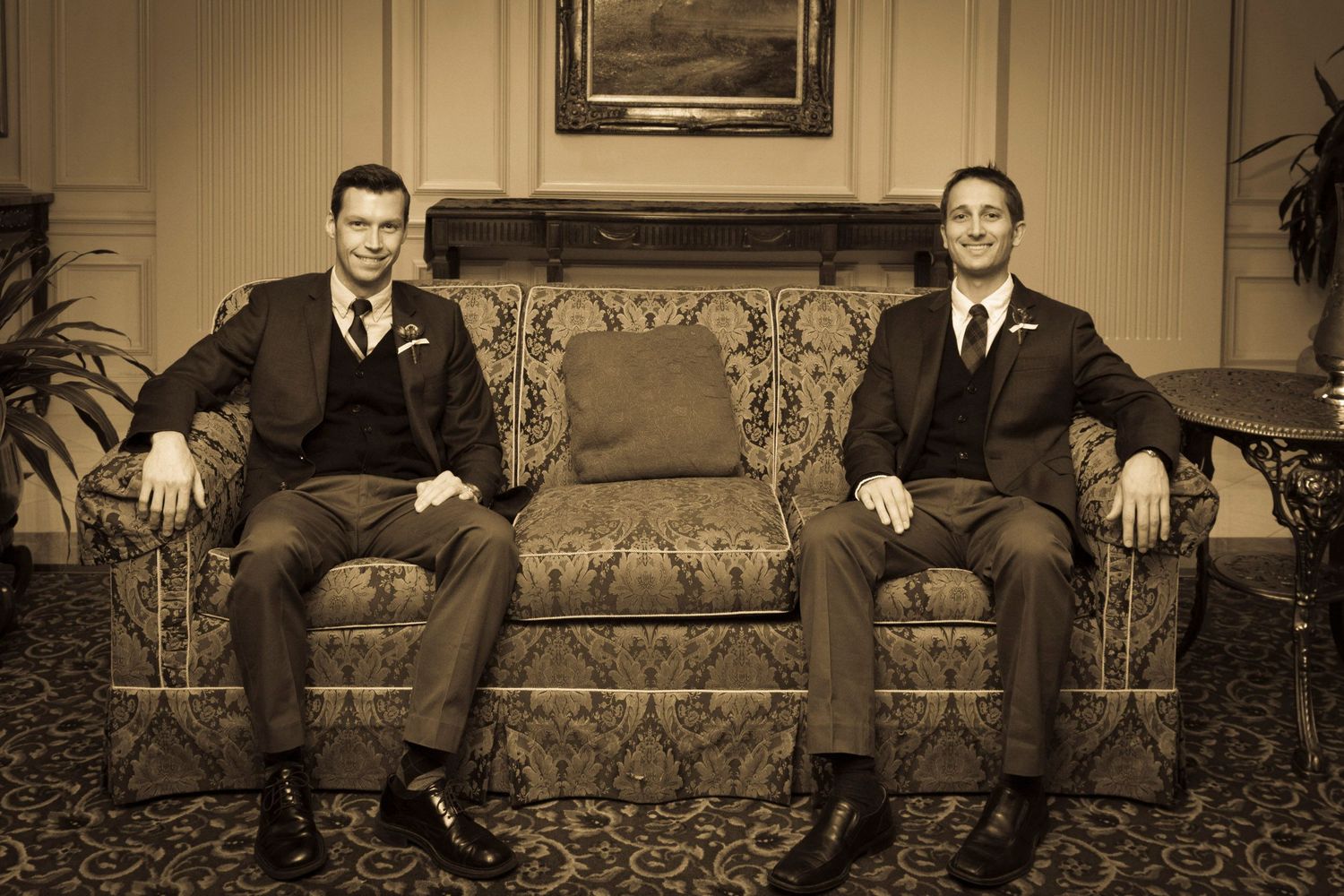 Two grooms in dark suits seated on an ornate sofa in a sepia-toned wedding portrait.