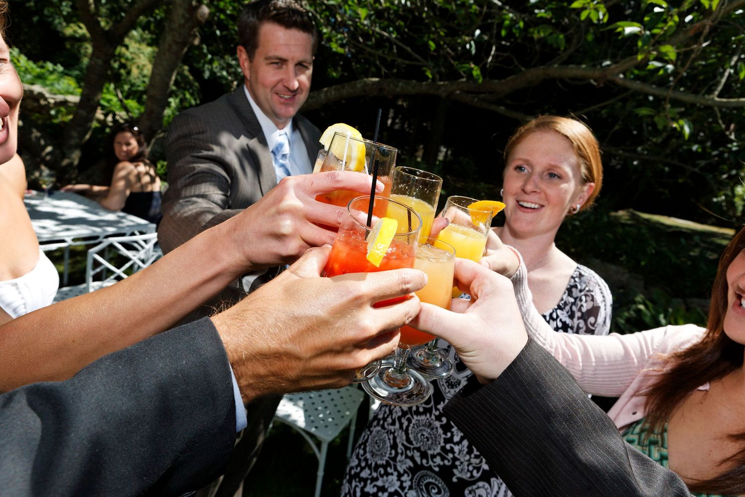 Wedding guests toasting with colorful cocktails outdoors under trees.